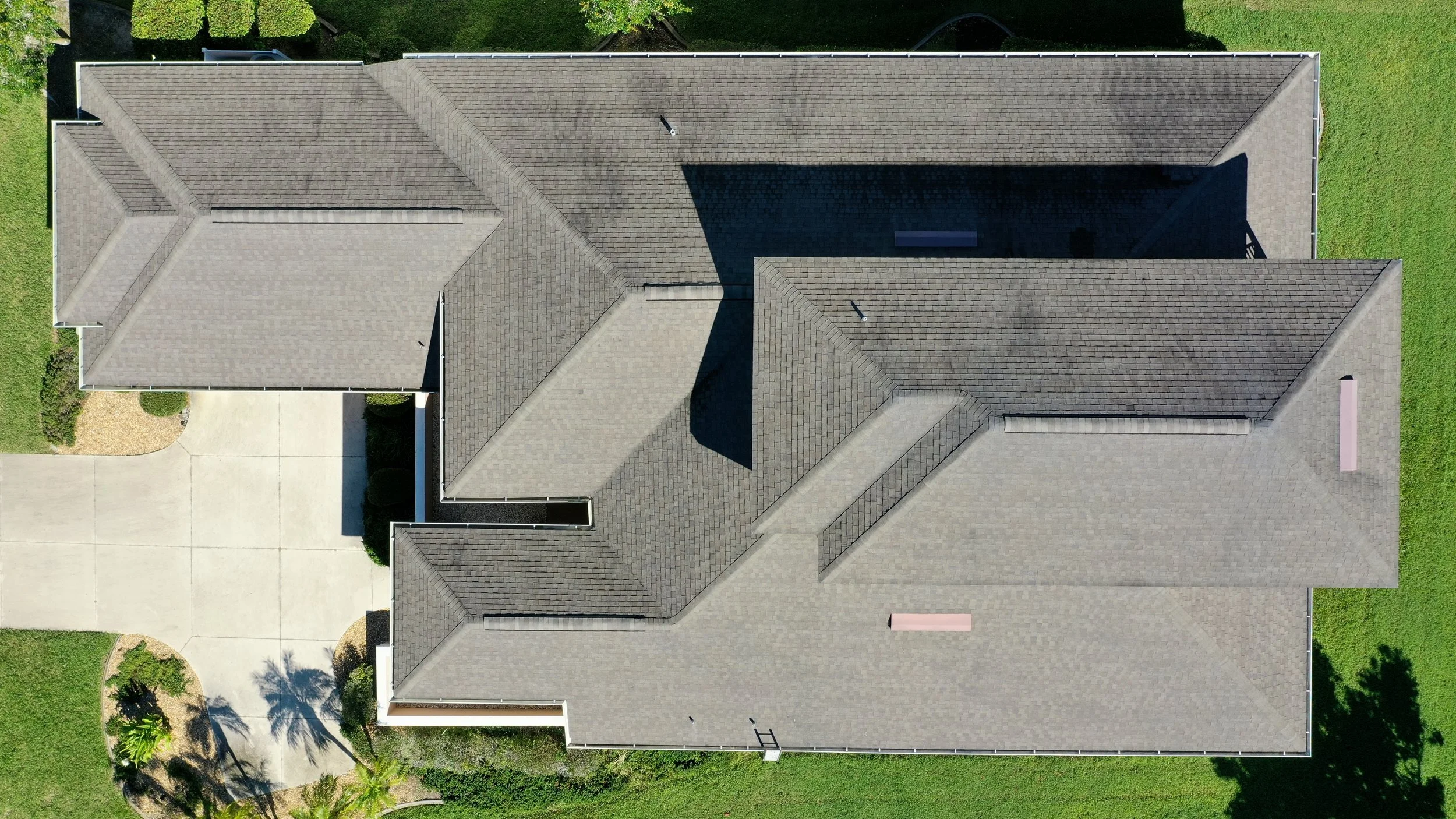 Aerial view of a house with multiple gray shingle roofs, a concrete driveway, and green lawn.