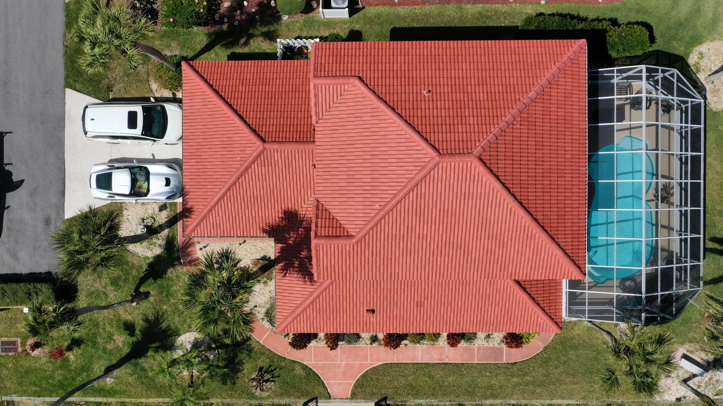 Aerial view of a house with a red tile roof, palm trees, a driveway with two white cars, and a screened-in swimming pool enclosure.