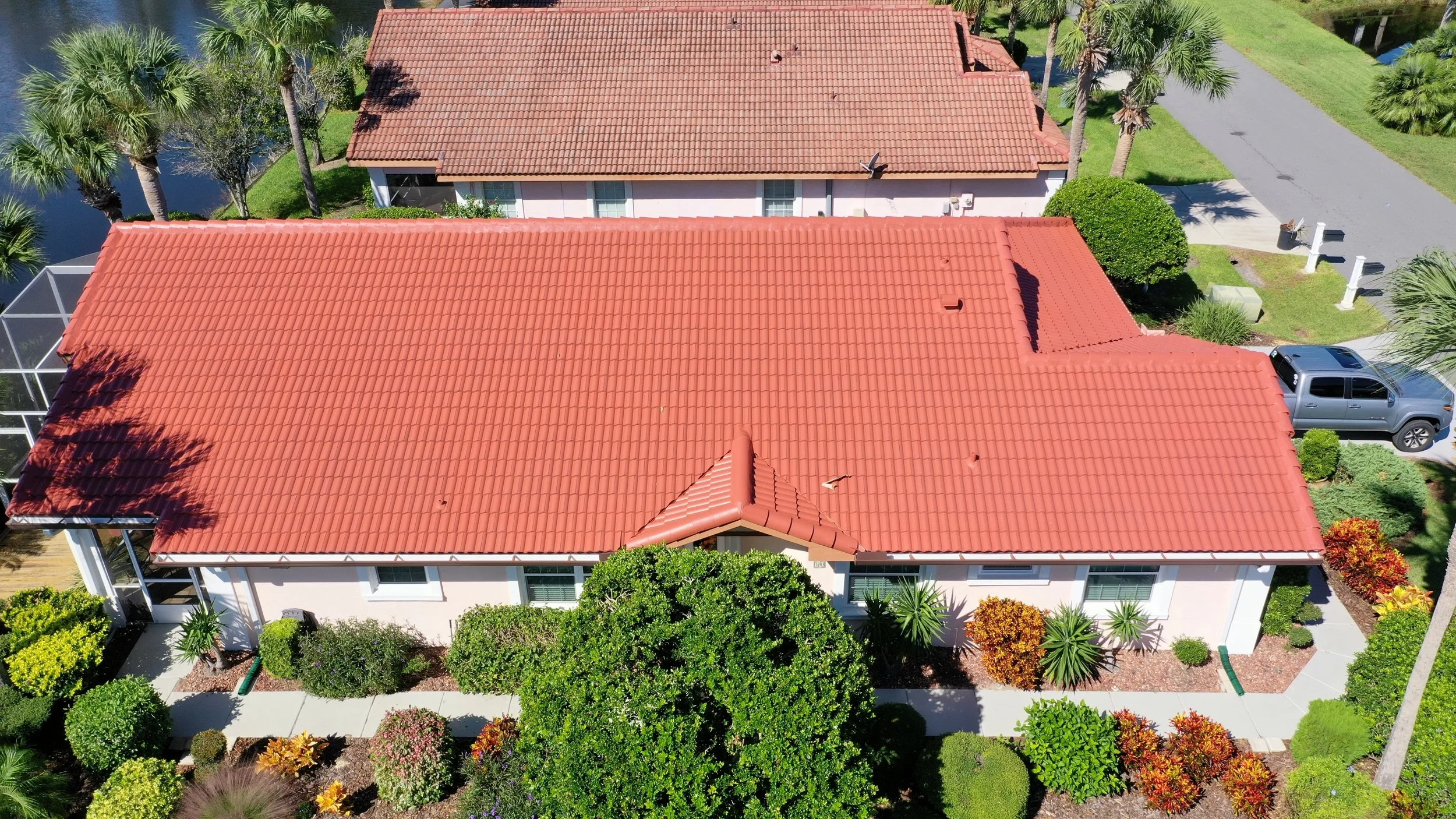 Aerial view of a residential house with a red tile roof, surrounded by green trees and manicured bushes, with a driveway and a parked truck.