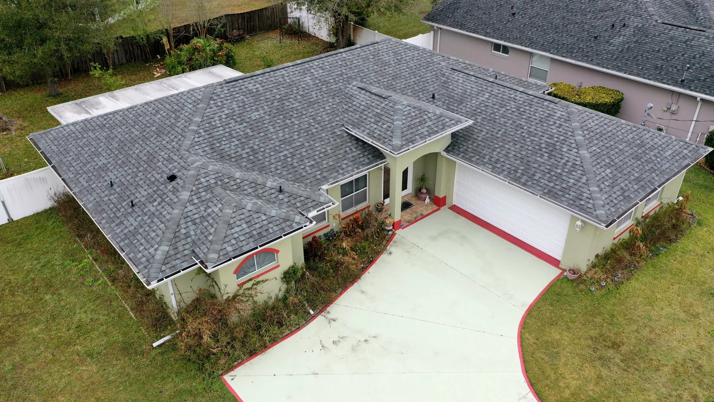 Aerial view of a single-story house with a also attached garage, a gray shingled roof, a driveway with red edging, and a small front yard with plants and shrubs, in a neighborhood with other houses and fenced backyard.