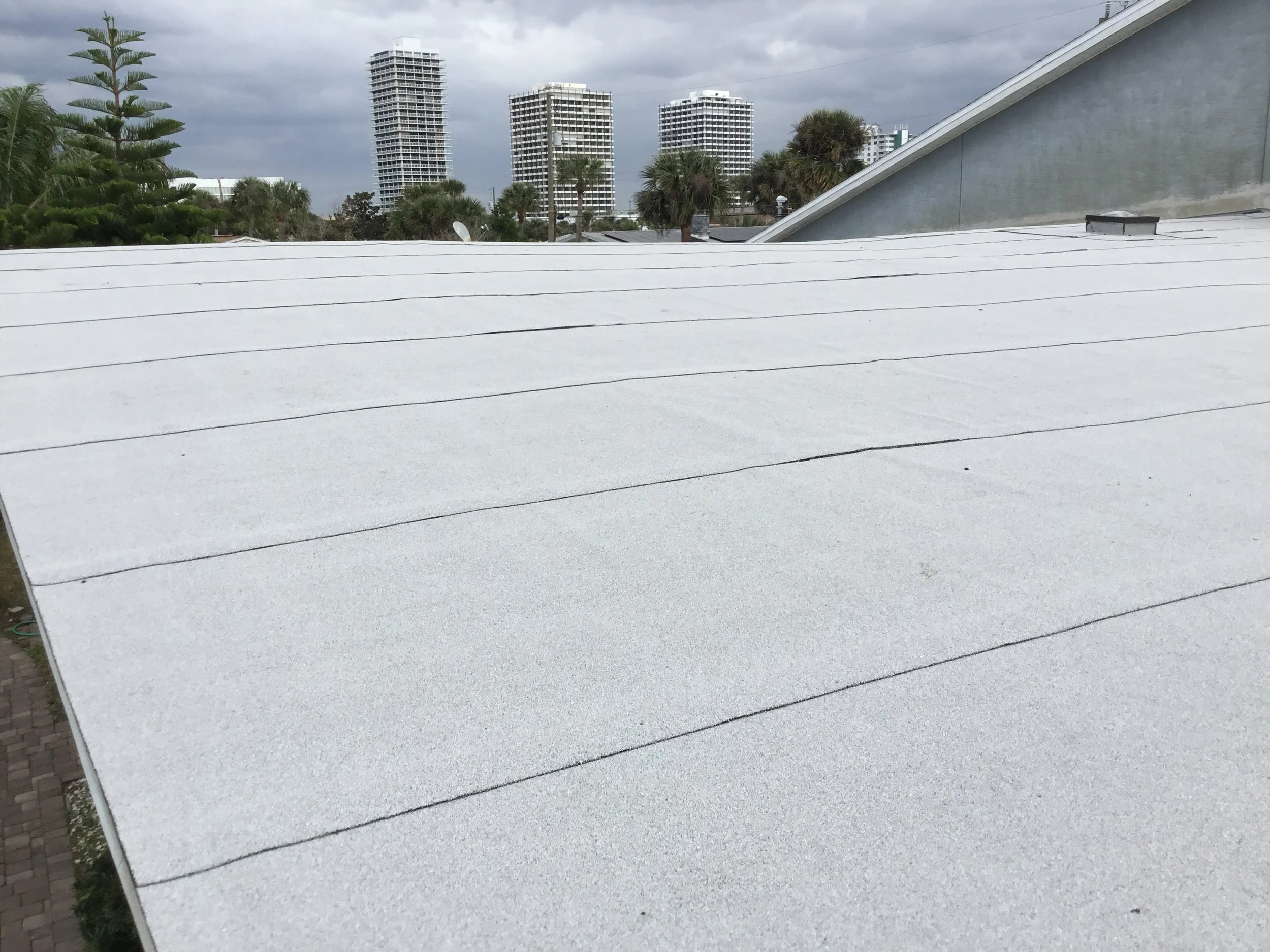 View of a white roof with tiles, city skyline with tall buildings, palm trees, and clouds in the background.