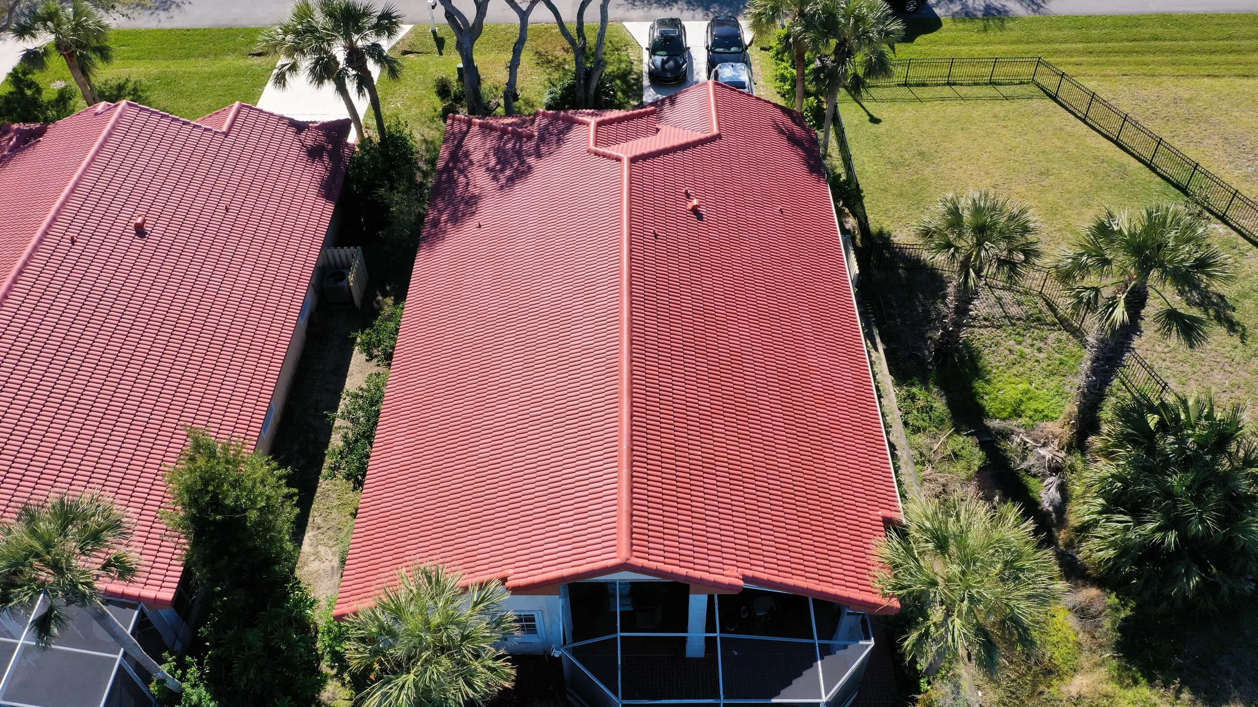 Aerial view of a house with a red tiled roof, surrounded by palm trees, a fenced yard, and a parking area with cars.