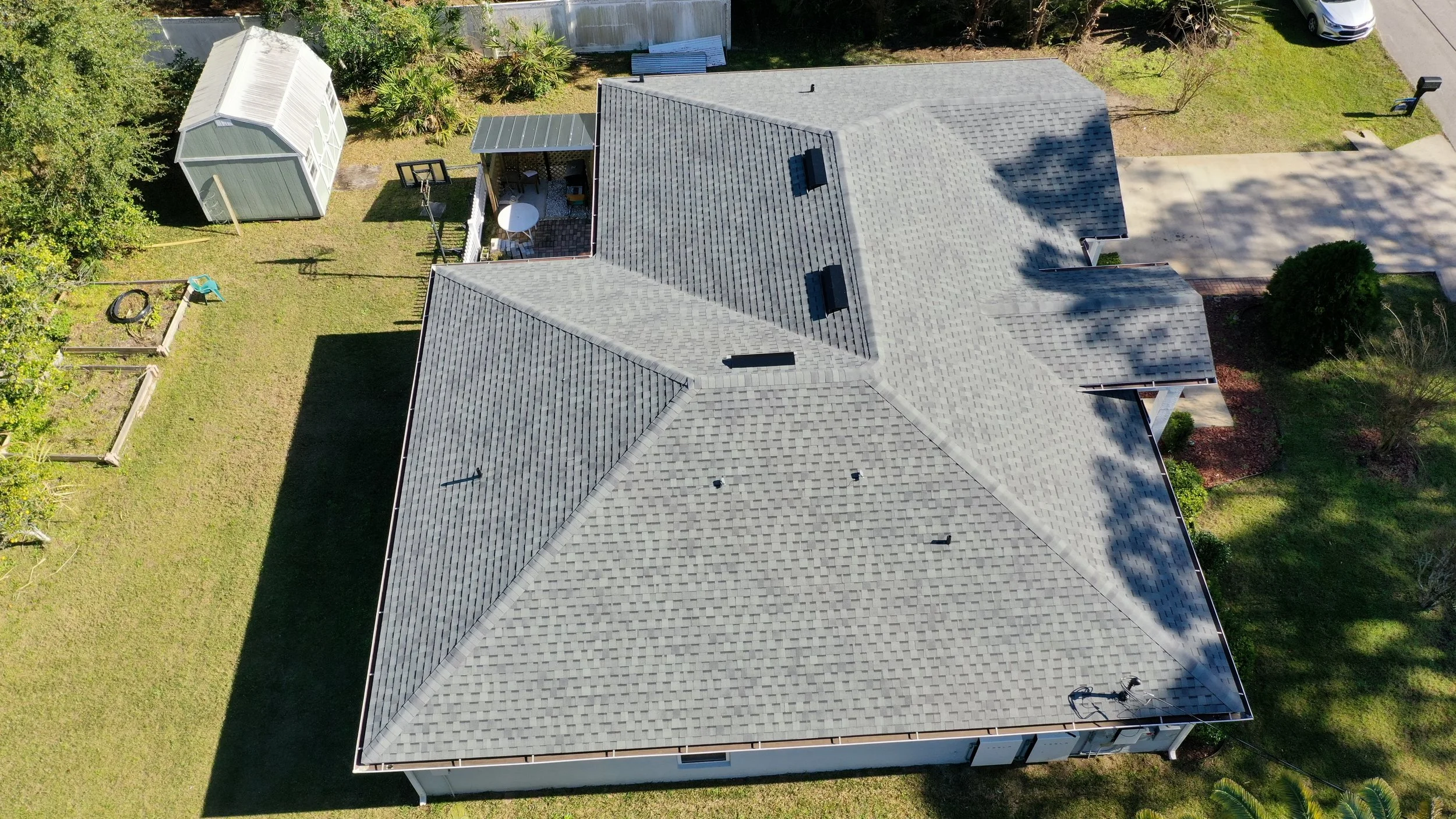 Aerial view of a house with a gray shingled roof, a backyard with a shed, a small patio with outdoor furniture, a basketball hoop, a black tire swing, a garden area, and a concrete driveway leading to a street.