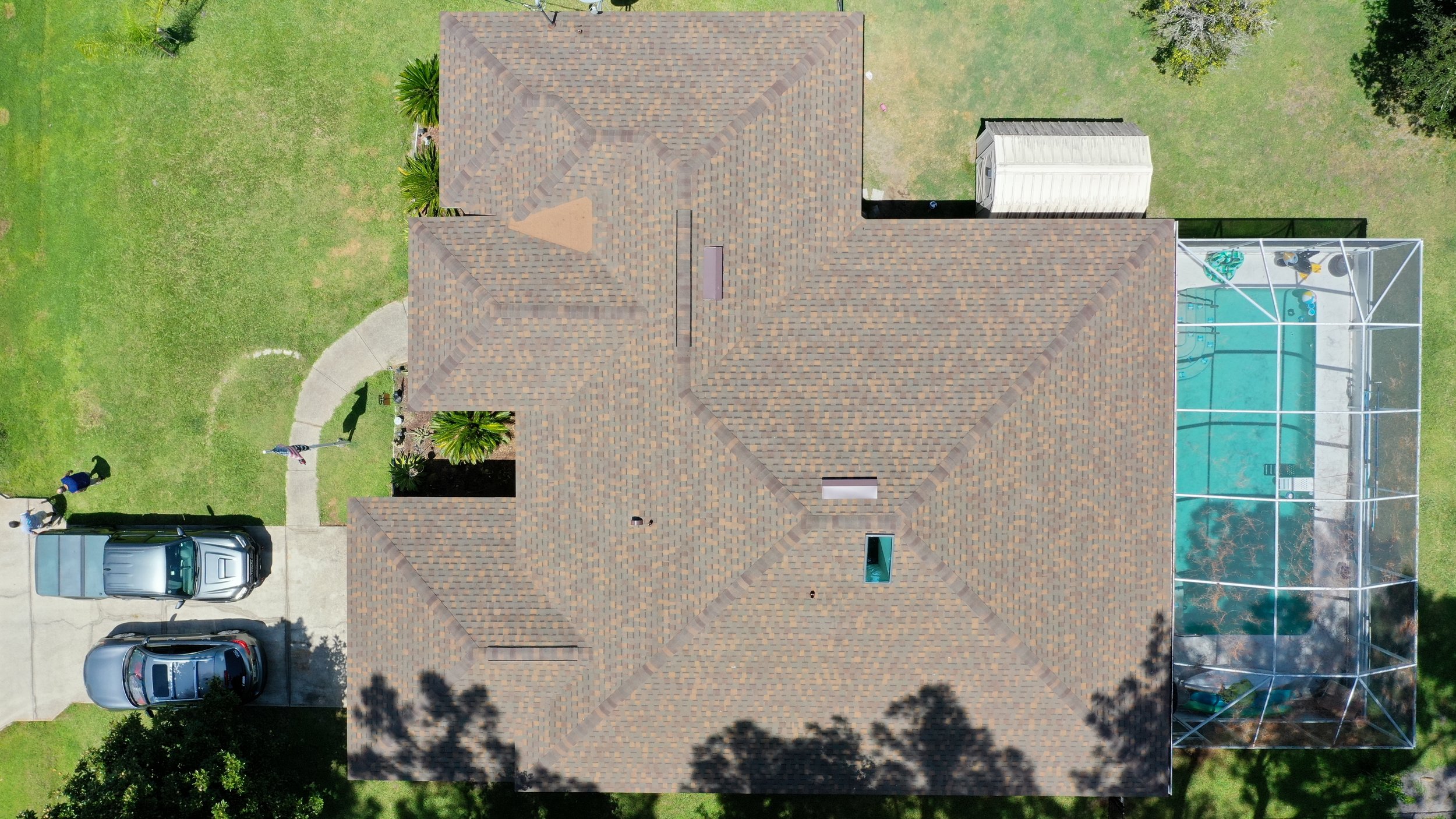 An aerial view of a house with a complex shingled roof, a driveway with two cars, a lawn with a curved walkway, and a screened-in pool enclosure on the right side.