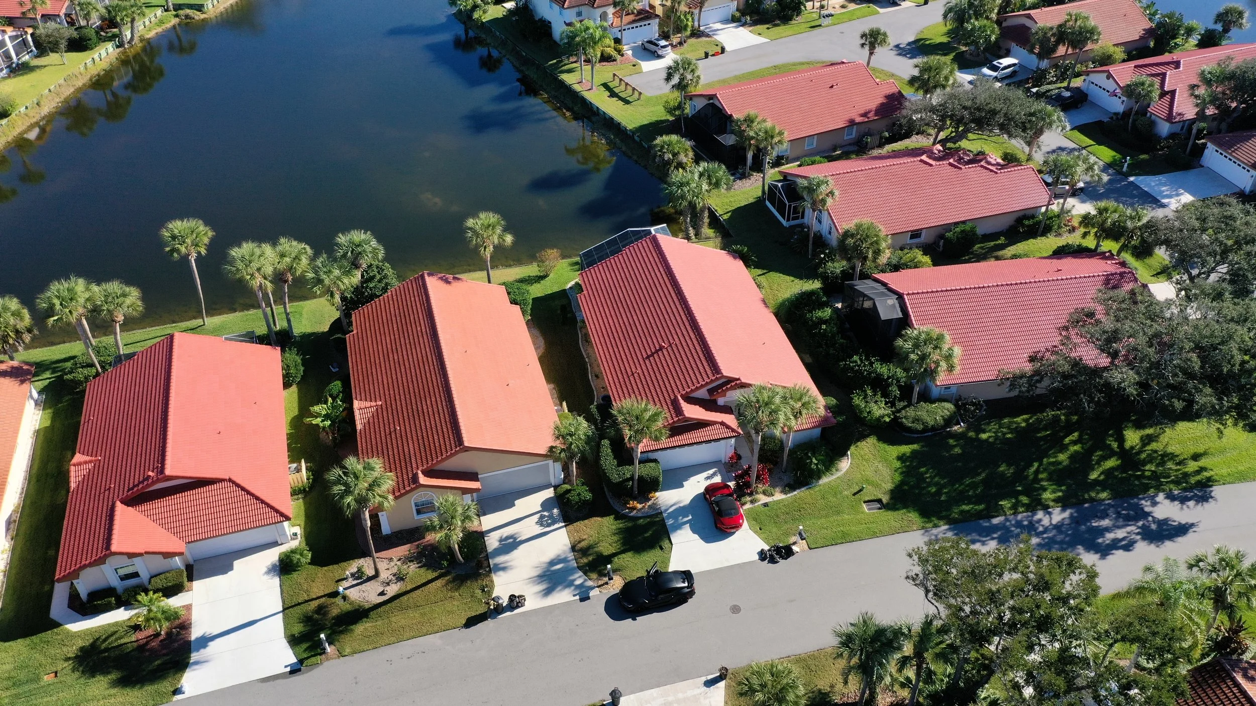 Aerial view of a residential neighborhood with houses featuring red tile roofs, green lawns, palm trees, and a pond nearby.