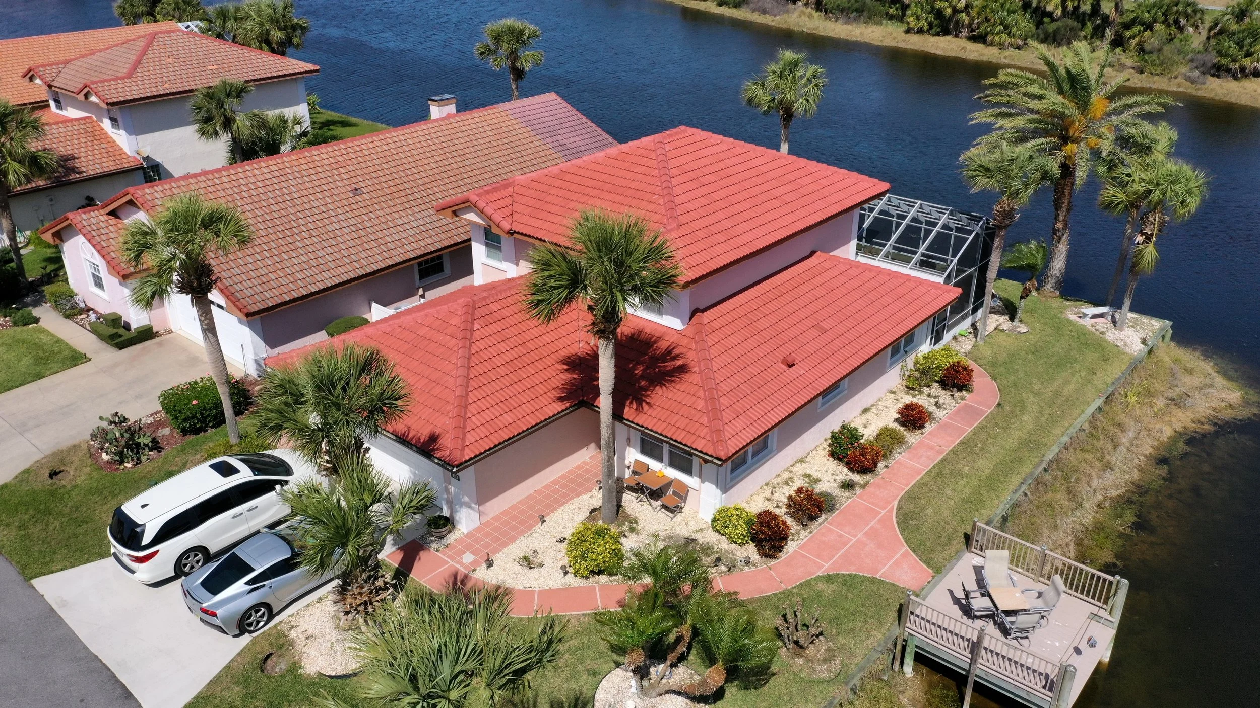 Aerial view of a residential house with a red-tiled roof, surrounded by palm trees, near a waterway. The property features a landscaped yard, a driveway with two parked cars, a small patio with outdoor furniture, and a screened porch area.