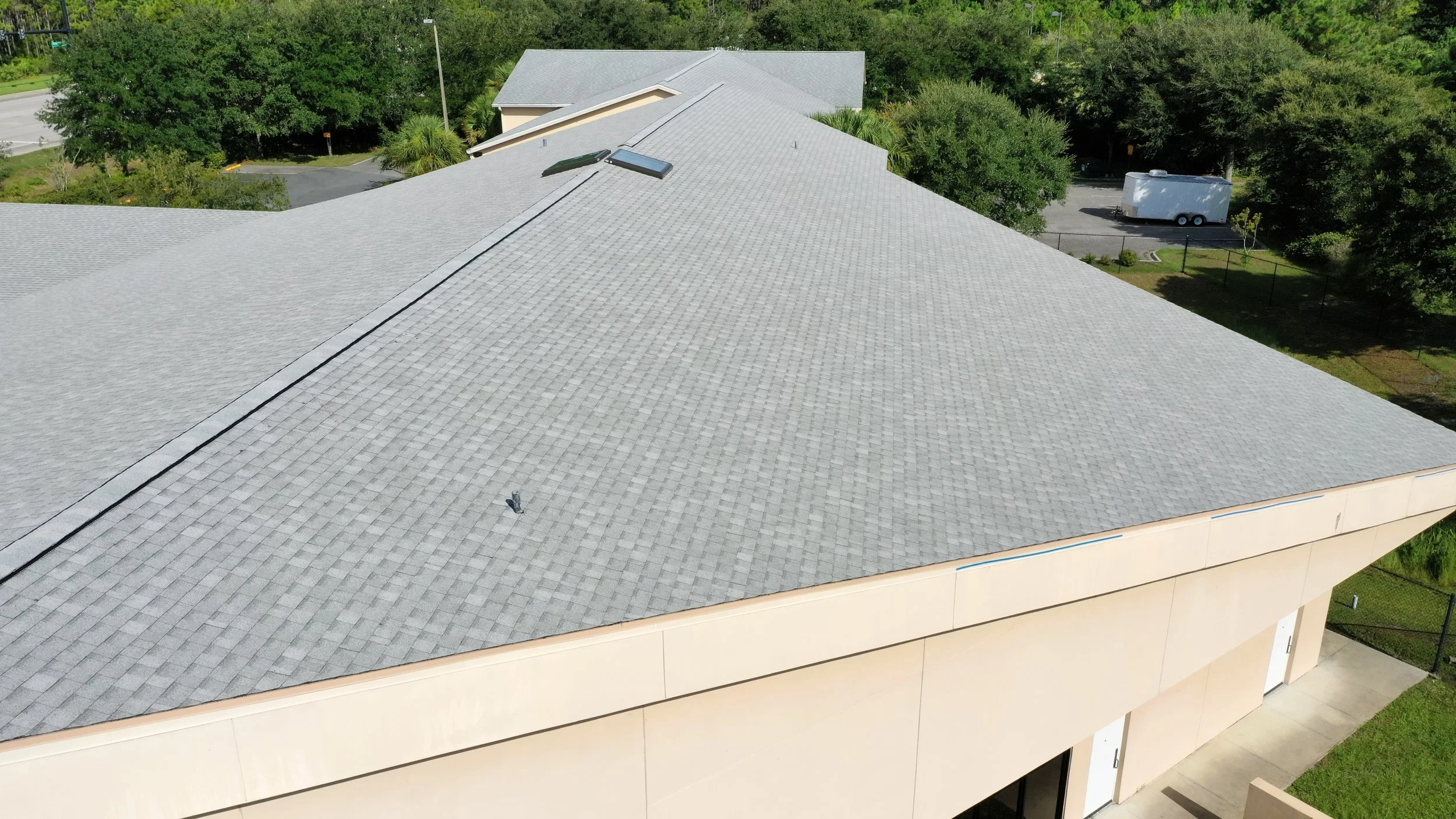 Aerial view of a large building with a gray shingle roof, surrounded by trees and parking lot, with a white trailer parked outside.