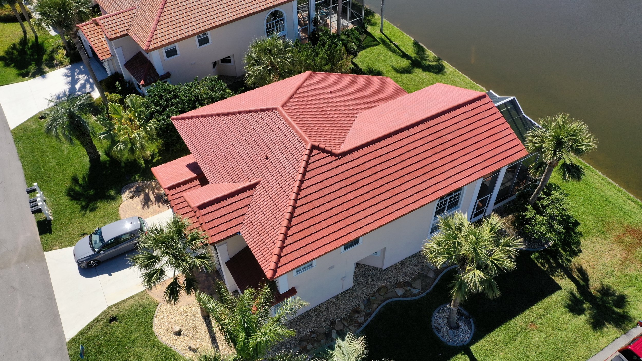Aerial view of a house with red tile roof, surrounded by a green lawn, palm trees, and a water body on the right.
