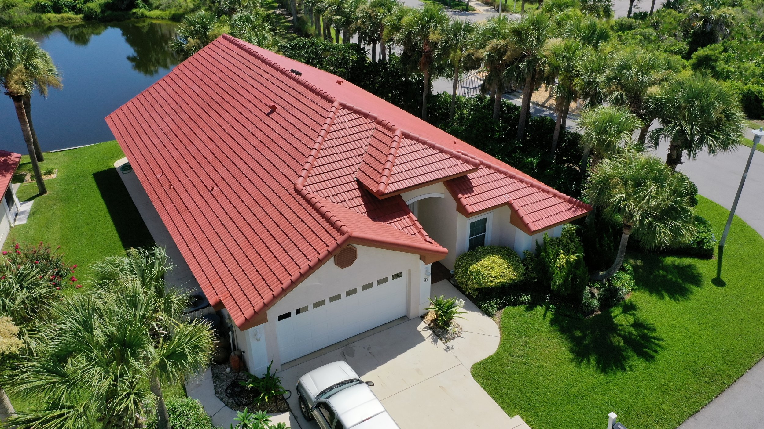 Aerial view of a house with a red tile roof, surrounded by green lawn, palm trees, and a pond nearby.