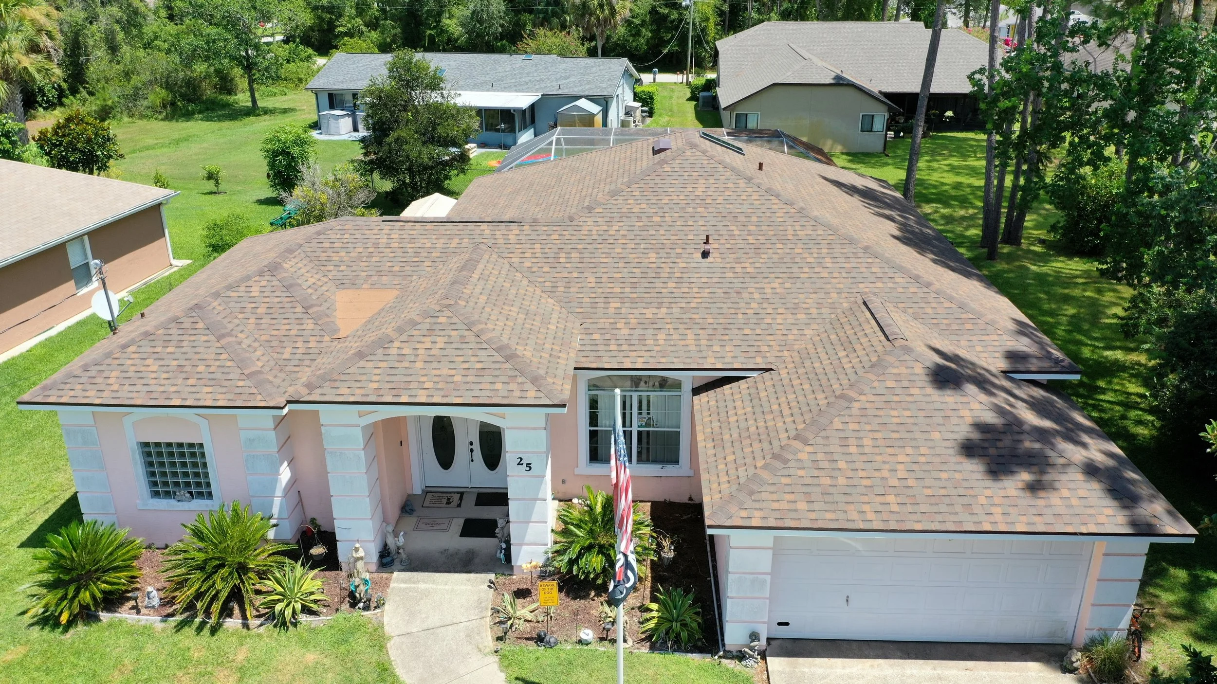 An aerial view of a suburban house with a brown shingle roof, pink exterior walls, a white garage door, and a front porch with decorative plants and garden statues. The yard has green grass, trees, and neighboring houses visible in the background.