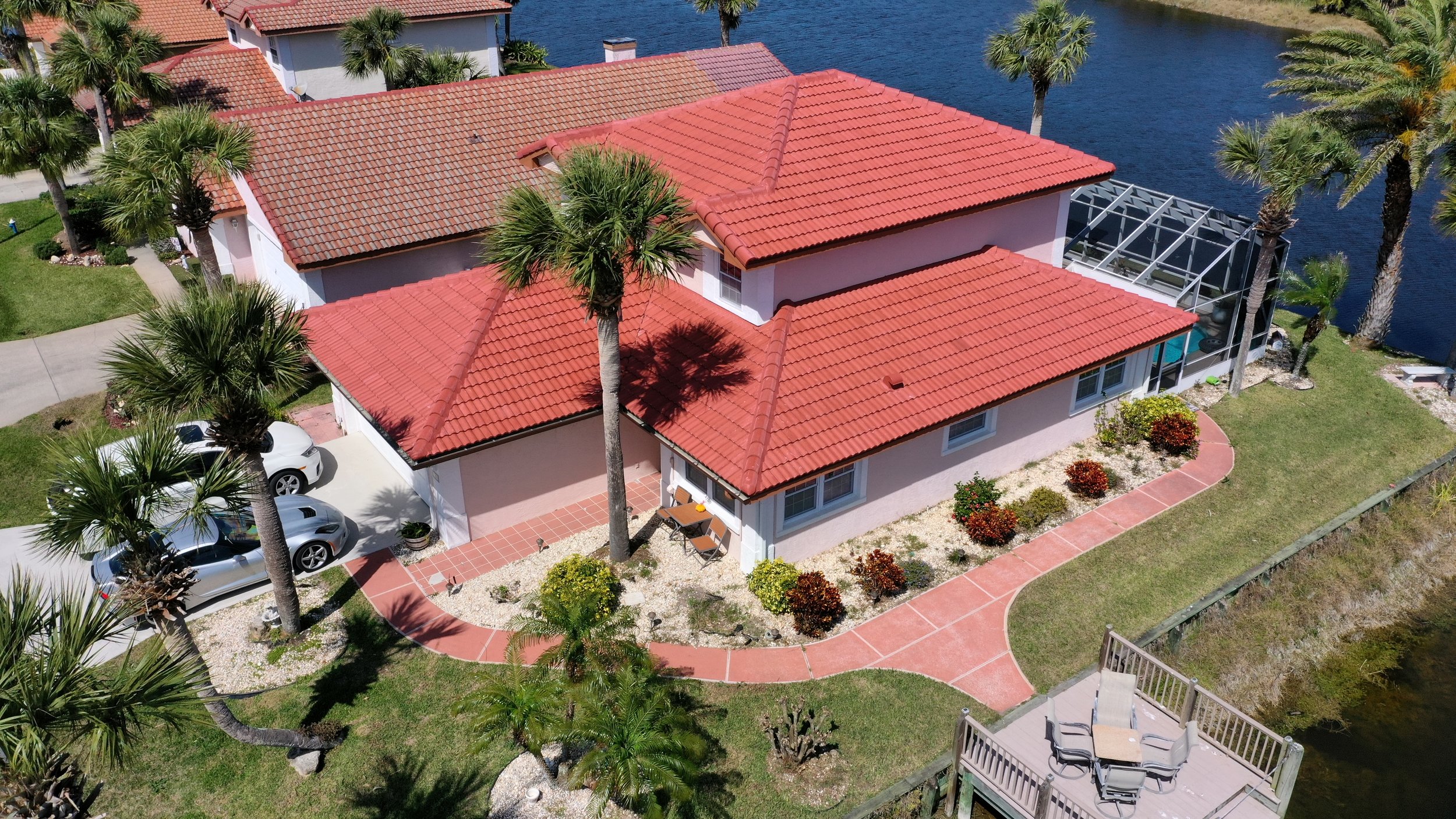 Aerial view of a house with red tile roof, surrounded by palm trees, parking area with cars, a waterfront yard with a dock, and a curved pathway.