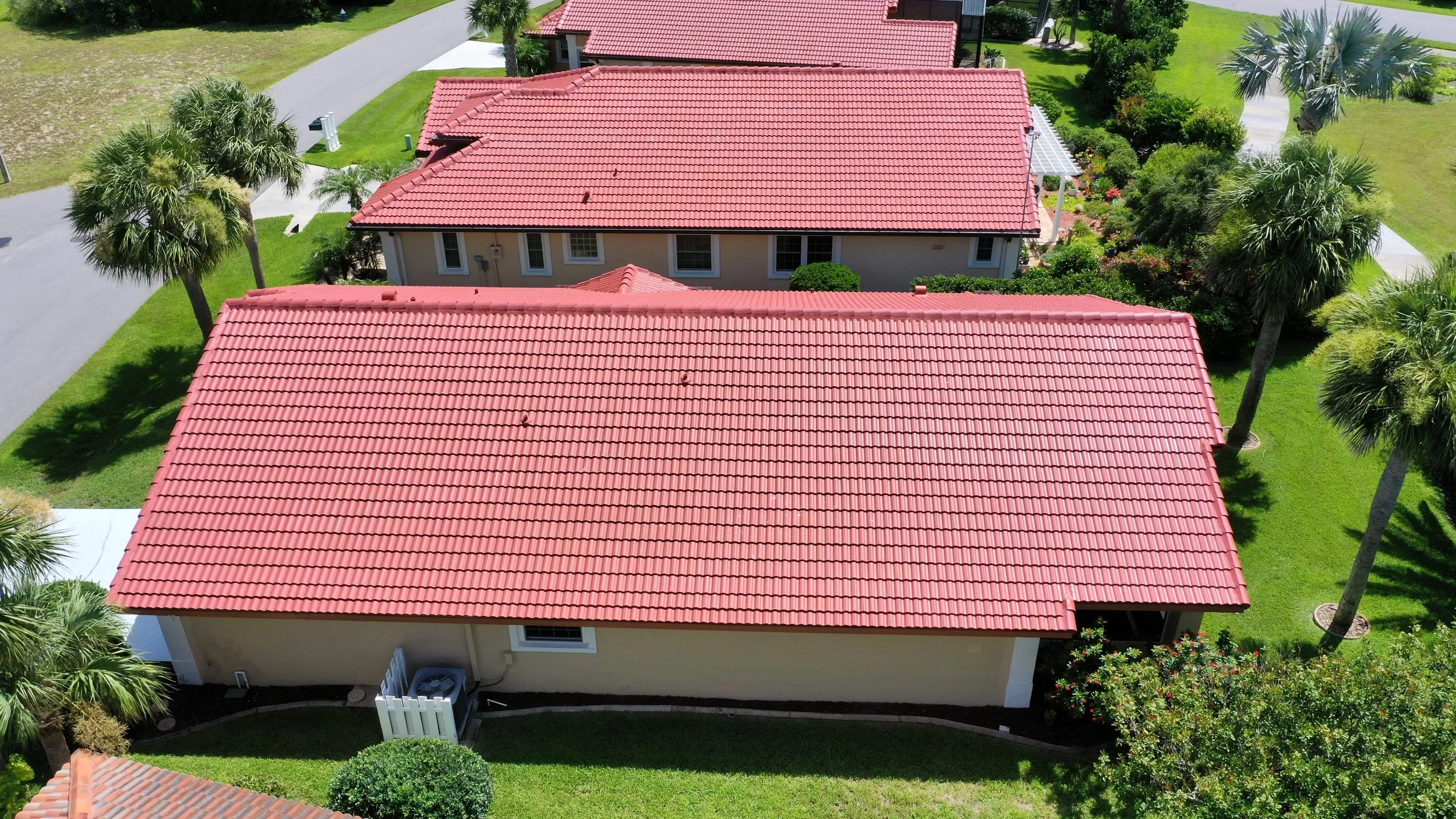 Aerial view of houses with red tile roofs, surrounded by green lawns and palm trees.