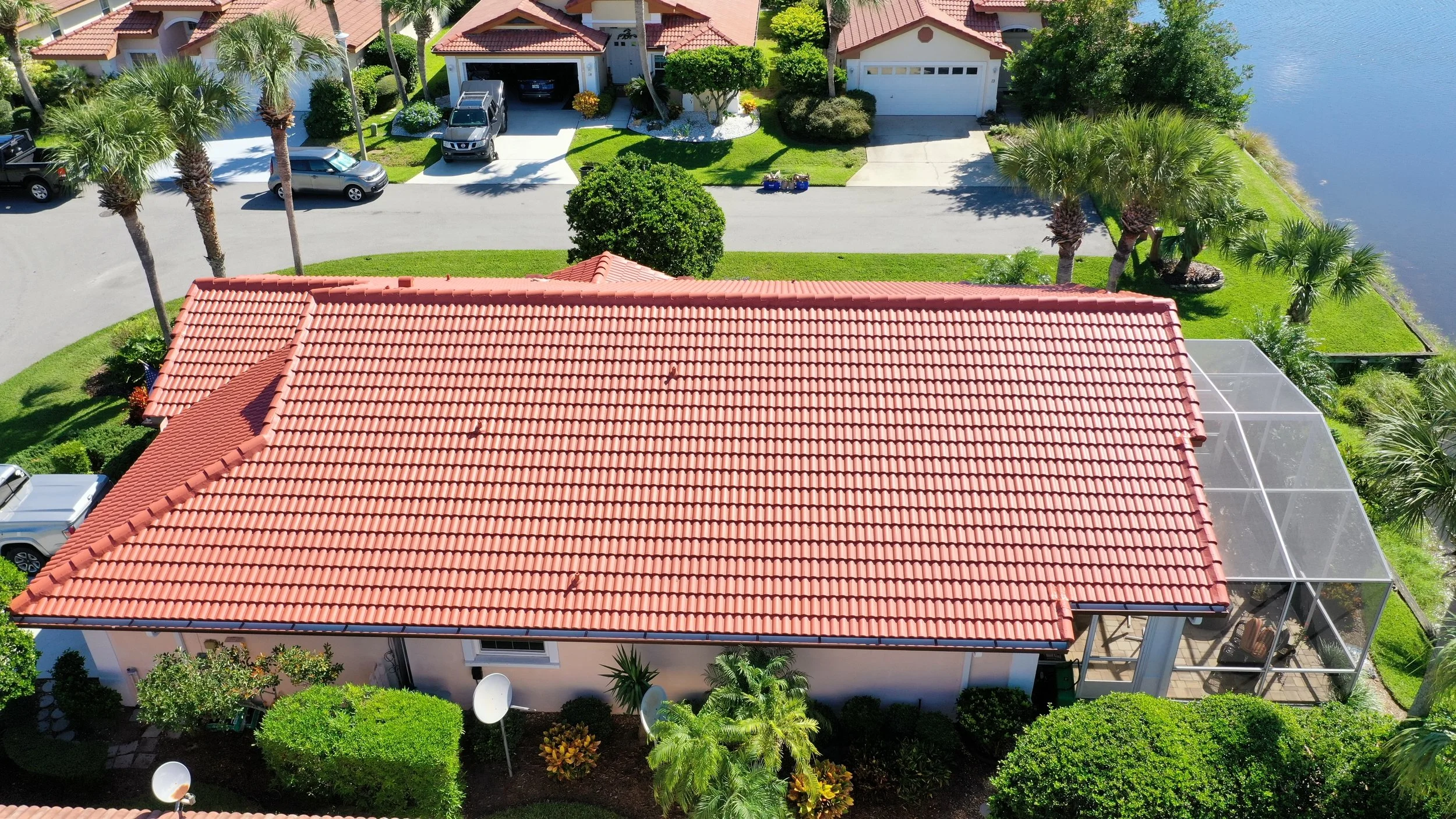 Aerial view of a house with a red-tiled roof, surrounded by trees and green lawns, with a screened porch on the right. Several cars are parked in driveways and along the street, with a body of water visible on the right side of the image.