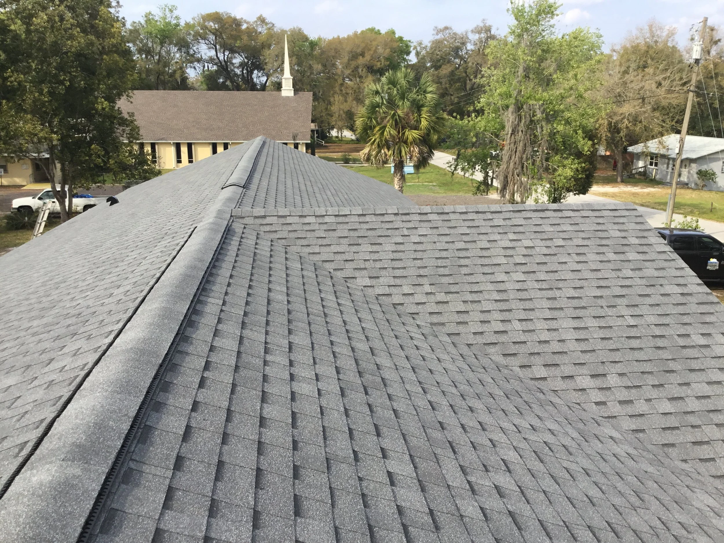 View of a gray shingled roof on a residential house with trees and a church in the background.