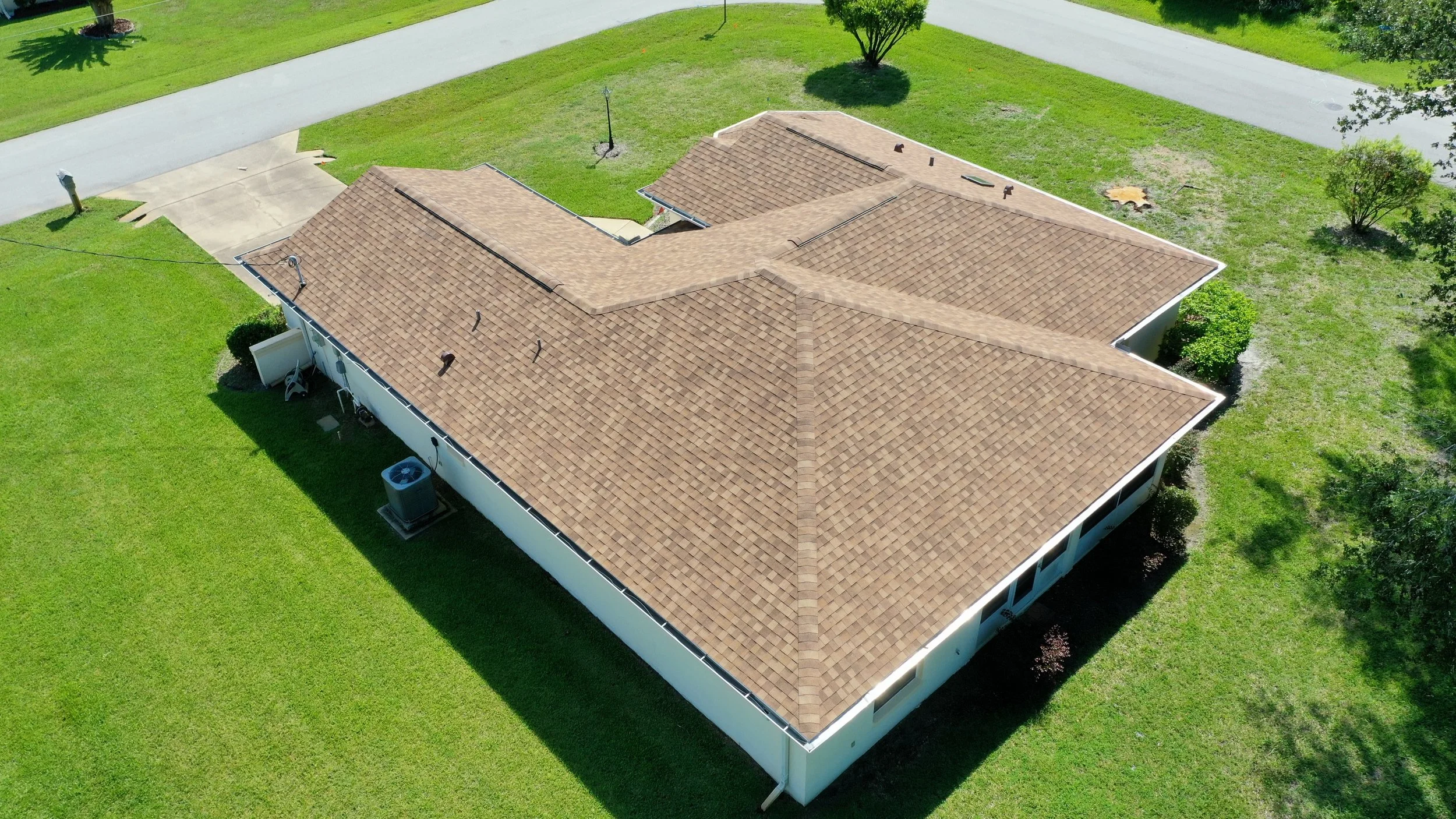 A residential house with a multi-section beige shingle roof, surrounded by a green lawn, driveway, and trees.