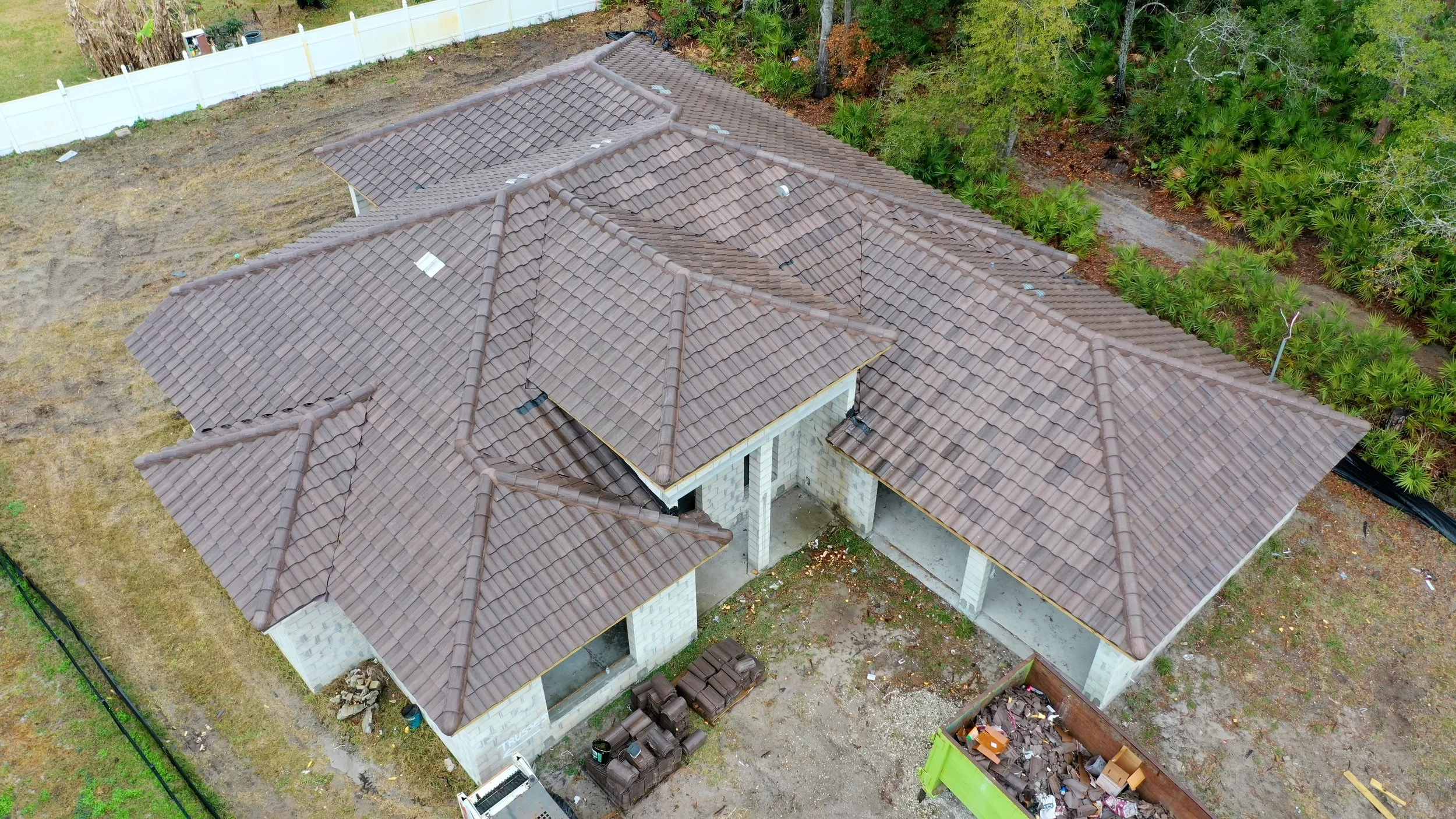 Aerial view of a house under construction with brown tiled roof, unfinished walls, and a surrounding yard with construction materials, debris, and a green dumpster. Foliage and trees are visible in the background.