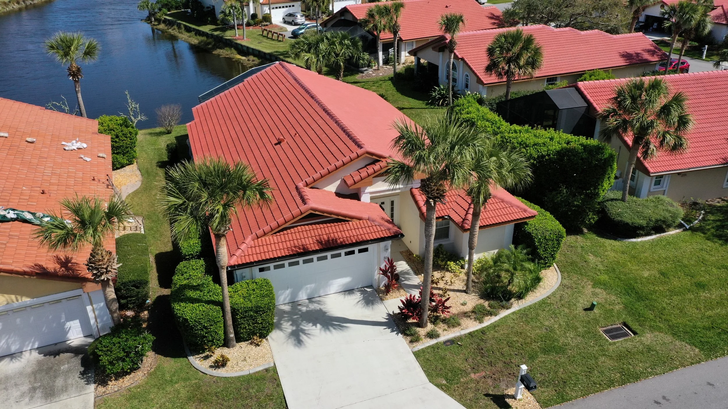 Aerial view of a house with a red tile roof, palm trees, a driveway, and a nearby waterway, with other similar houses in the neighborhood.