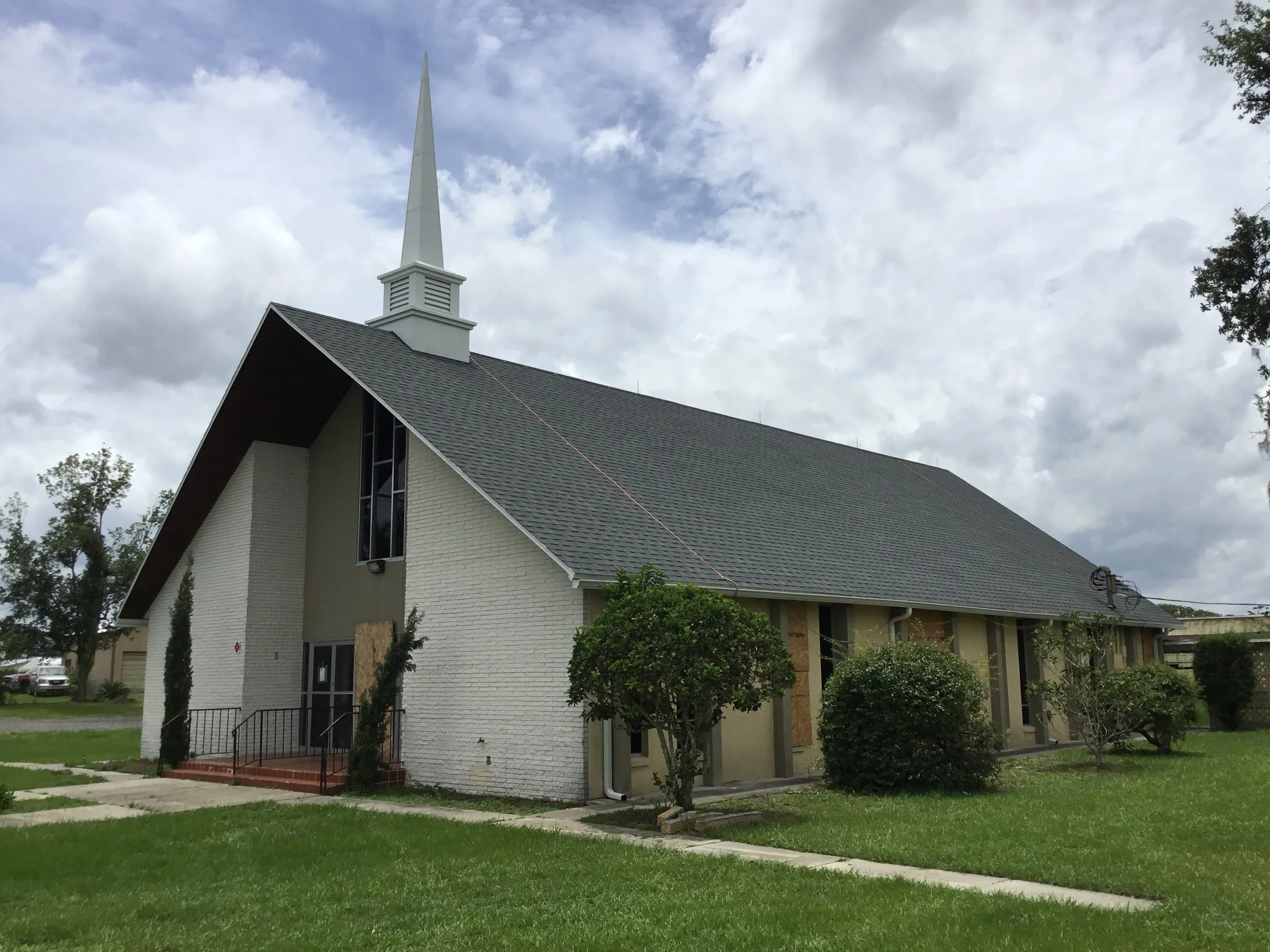 A church building with a steeply pitched gray roof, white brick walls, and a tall white steeple, surrounded by grassy lawn, trees, and bushes under a partly cloudy sky.
