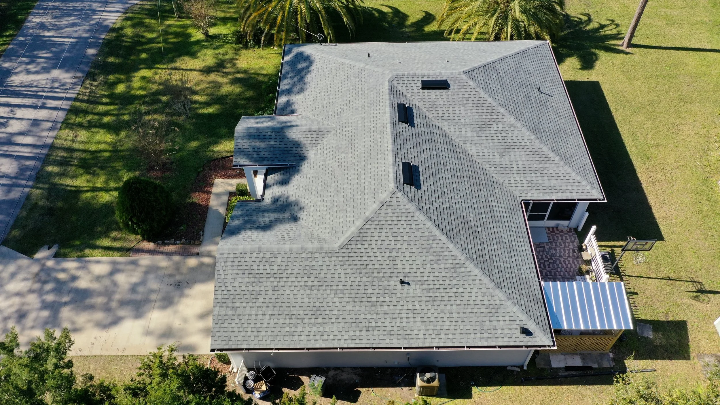 Aerial view of a house with a gray shingle roof, surrounded by green lawns, trees, and a driveway.