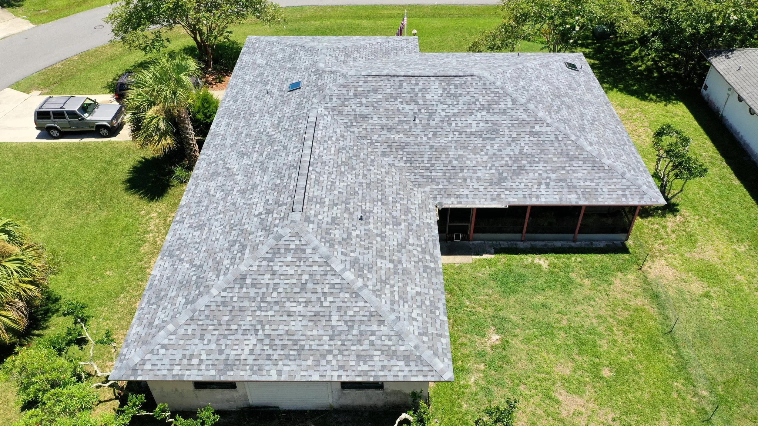 Aerial view of a house with a gray shingled roof, surrounded by green lawn and trees, with a driveway and parked vehicles.