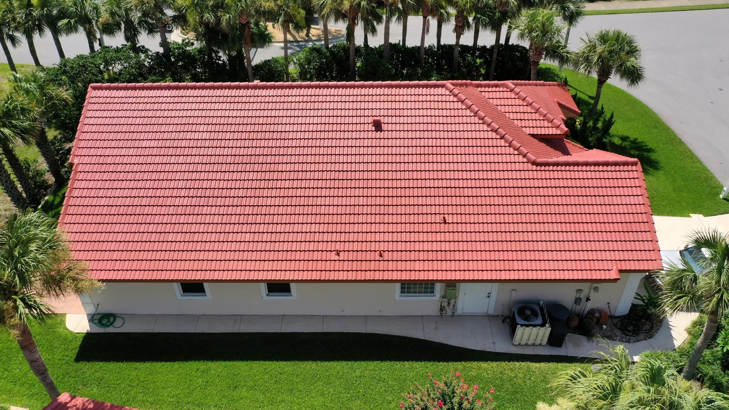 Aerial view of a beige house with a red tiled roof, surrounded by green lawn, palm trees, and a paved road.