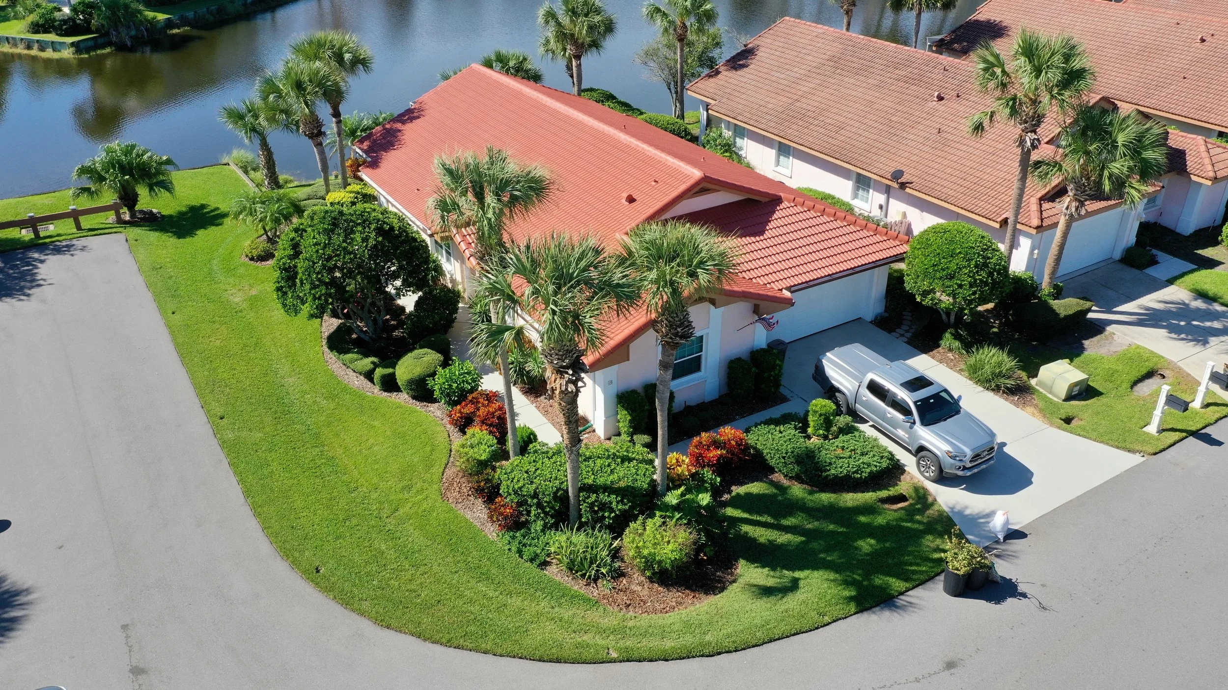 Aerial view of a house with a red tile roof, surrounded by landscaped yard with palm trees and bushes, near a body of water, with a driveway and parked silver truck.