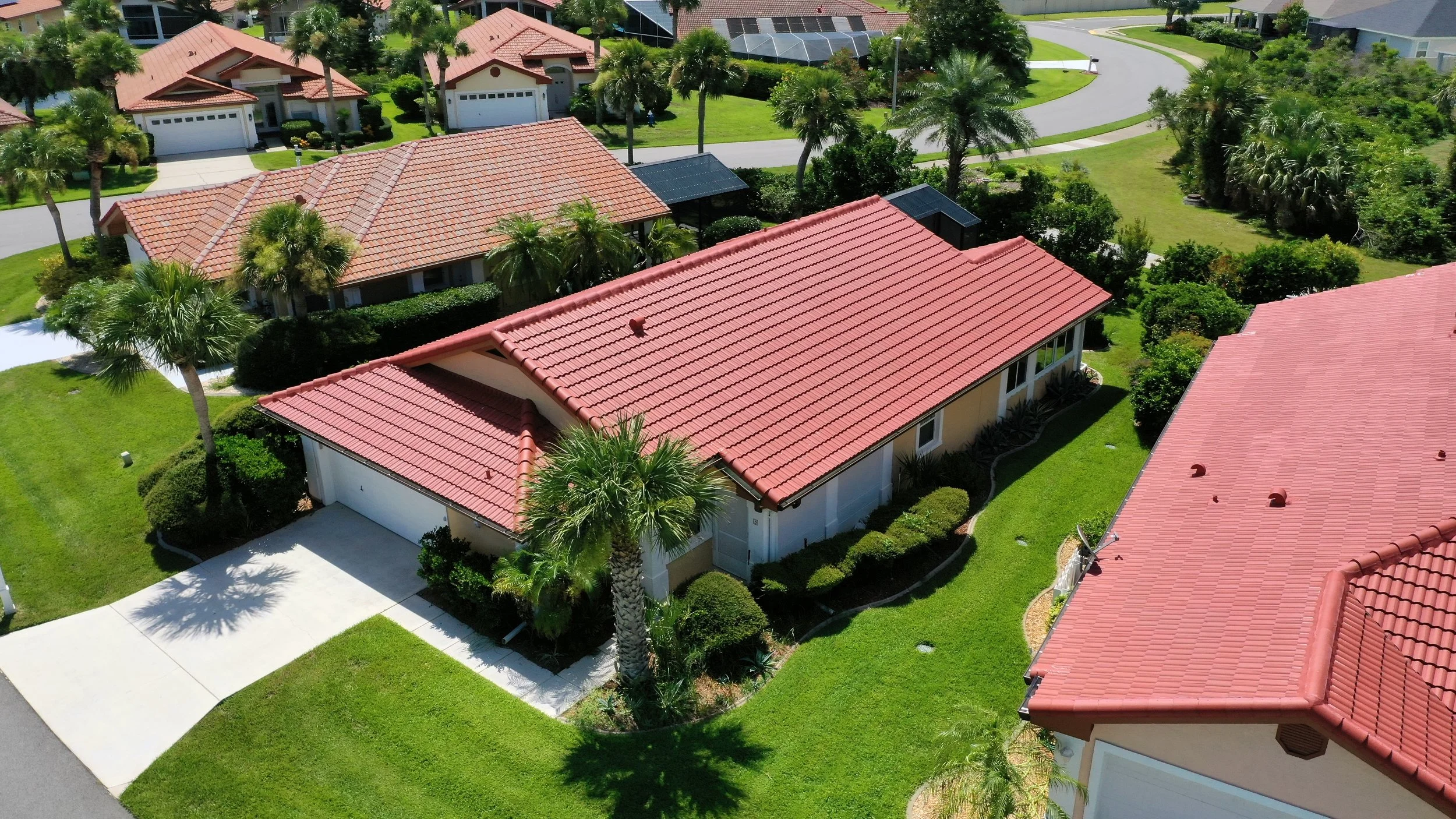 Aerial view of a suburban neighborhood with several houses featuring red tiled roofs, manicured lawns, and palm trees.
