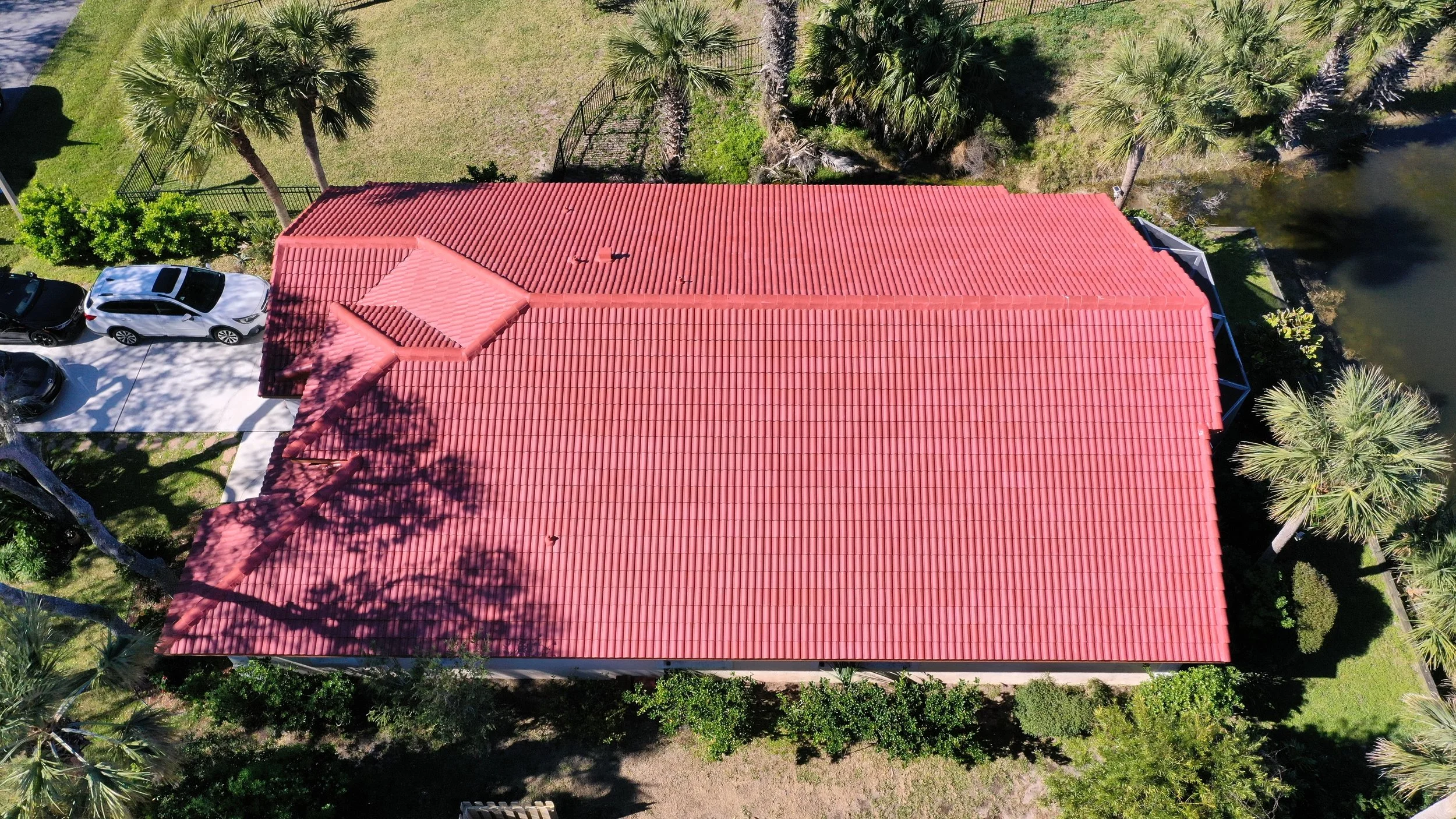 Aerial view of a house with a red tile roof, surrounded by palm trees, a small pond, and parked cars.