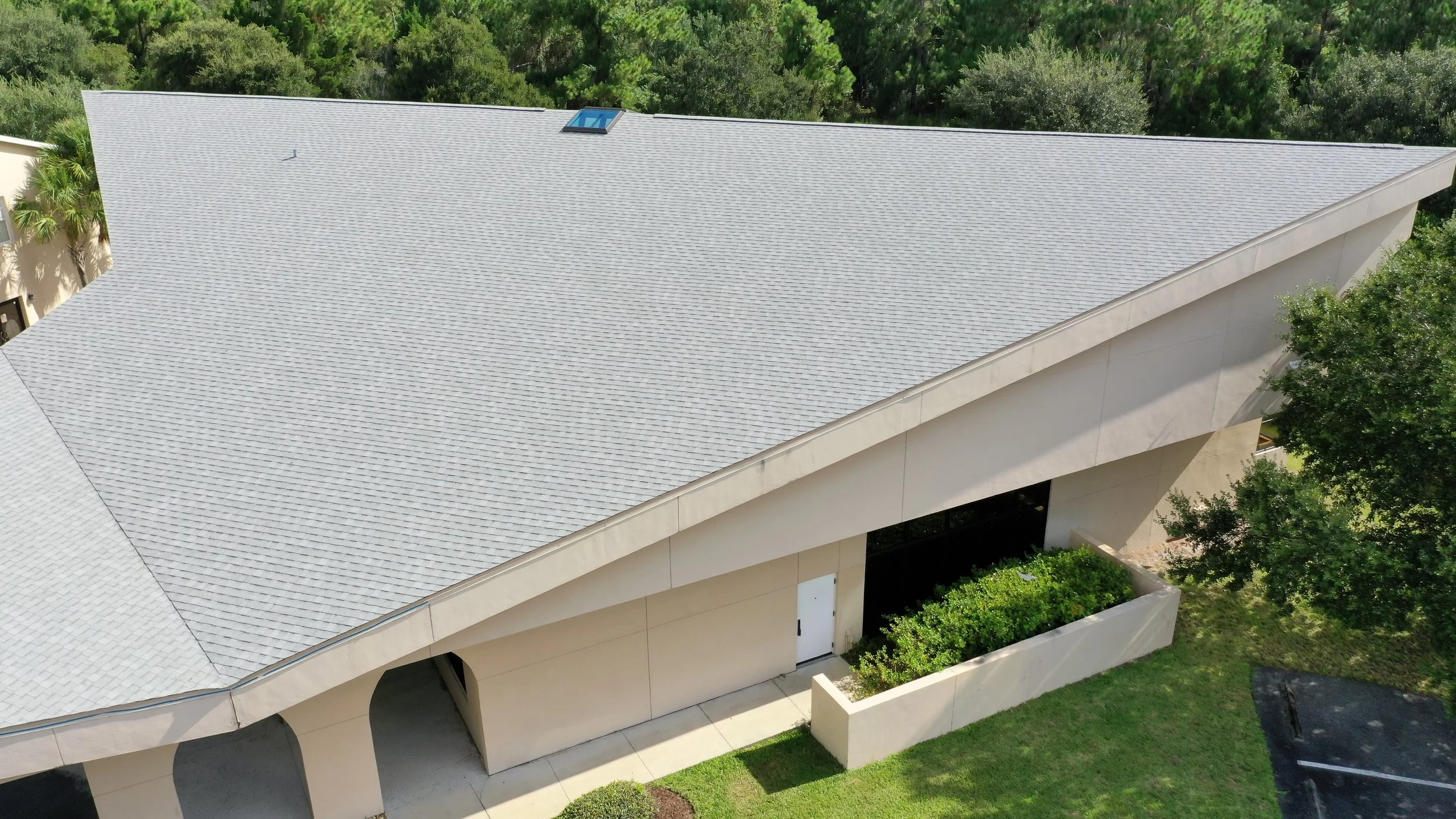 Aerial view of a modern building with a gray, shingle roof, surrounded by green trees and landscaped lawn, with a parking lot visible at the bottom right.