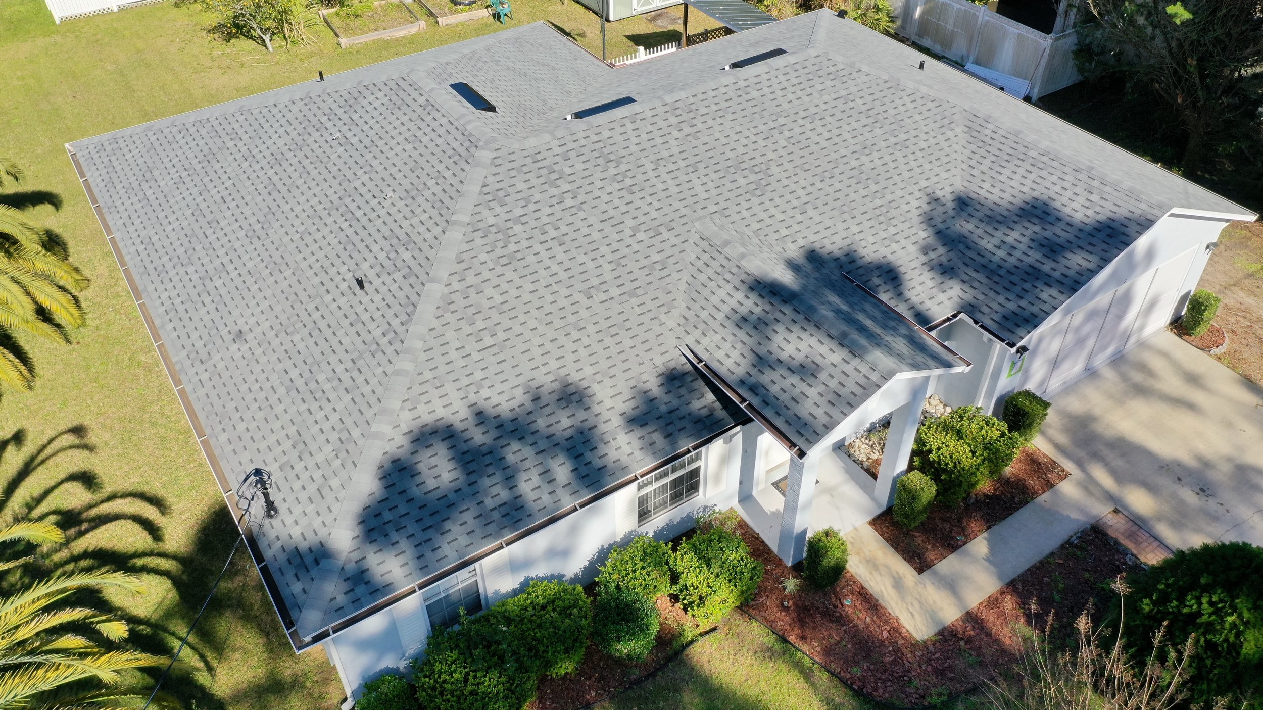 Aerial view of a house with a gray shingle roof, surrounded by green bushes, trees, and a paved driveway.