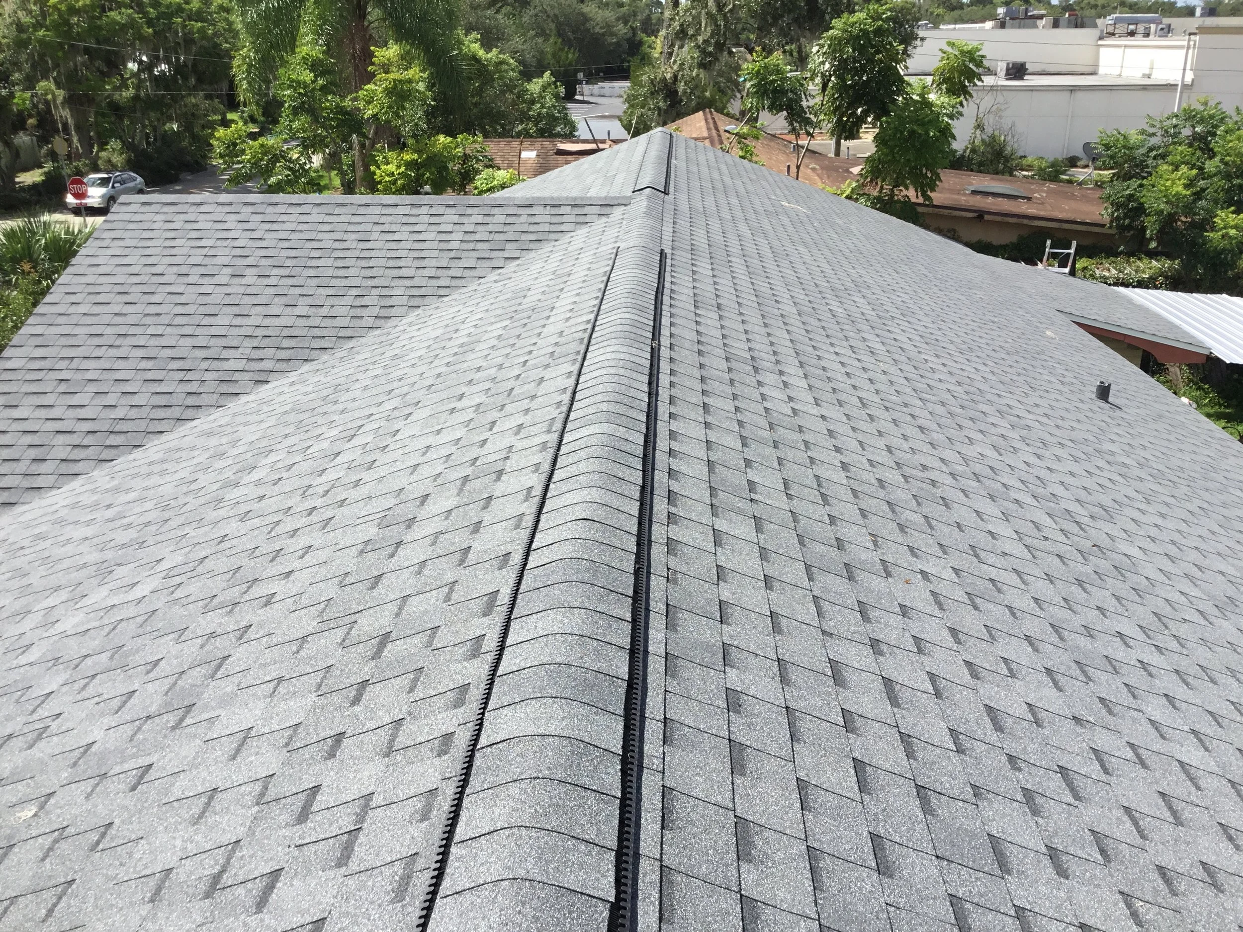 View of a residential roof with gray asphalt shingles, ridge vent along the peak, surrounded by trees and neighboring houses.