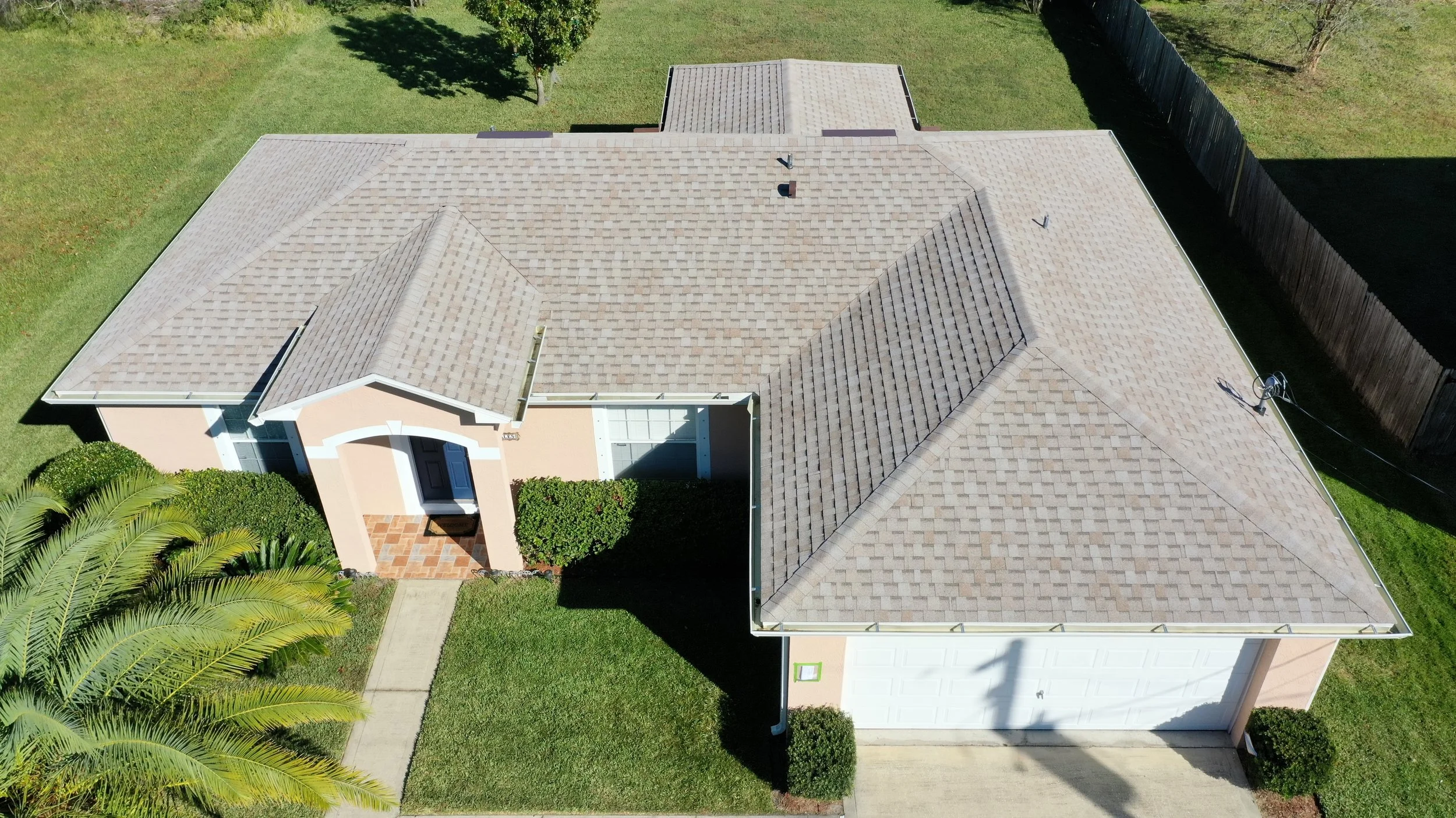 An aerial view of a house with a tan roof, white garage door, front door, and small front yard with trees, bushes, and a sidewalk.
