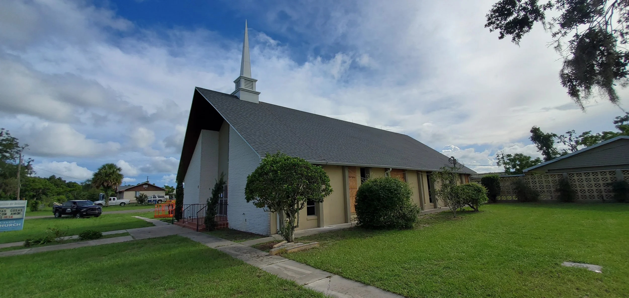 A church with a white brick exterior, gray roof, and tall steeple surrounded by a green lawn and trees under a partly cloudy sky.