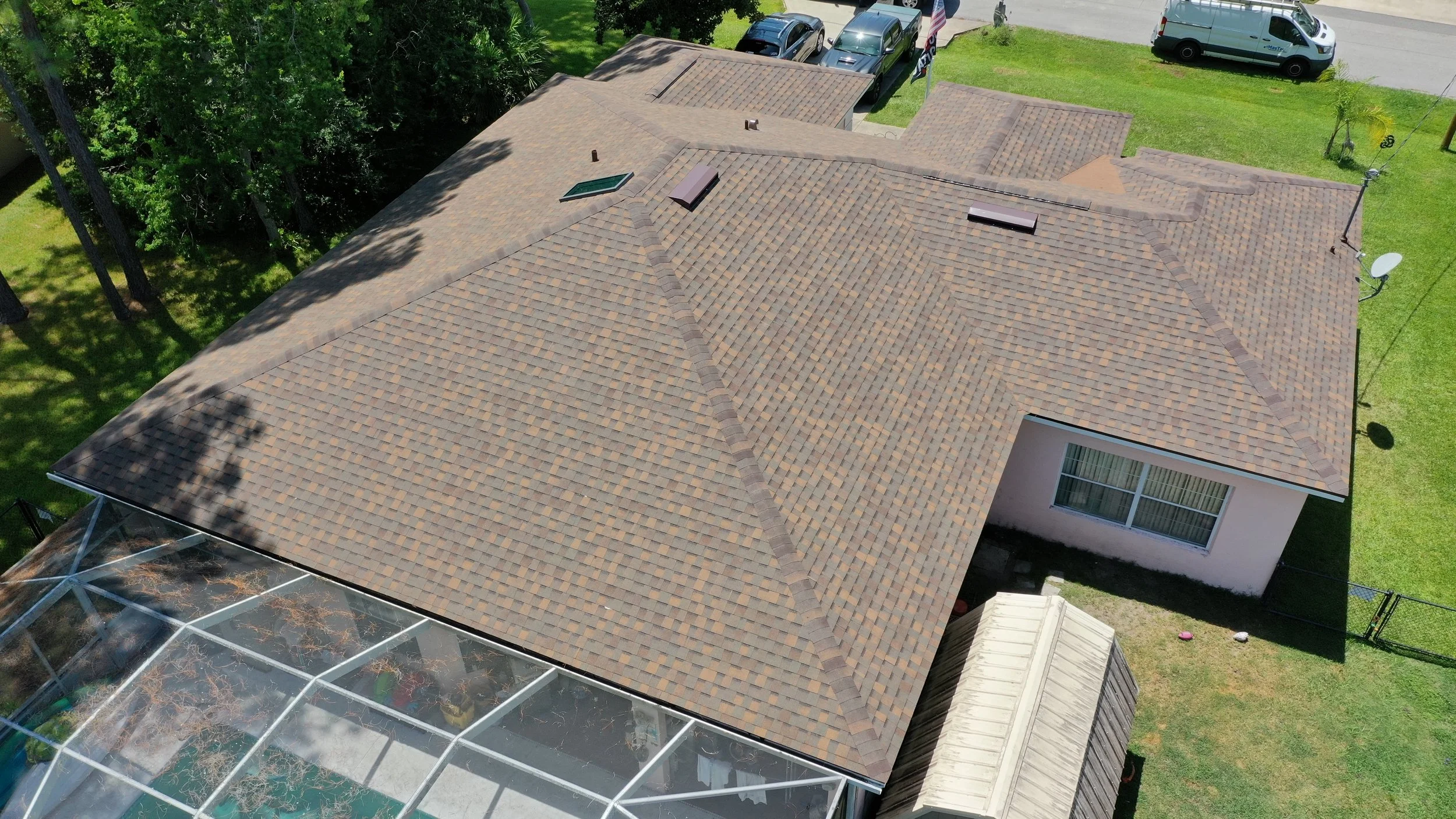 Aerial view of a house's roof with brown shingles, a skylight, and skylight vents, surrounded by green trees, a fenced yard, parked cars, and a satellite dish.