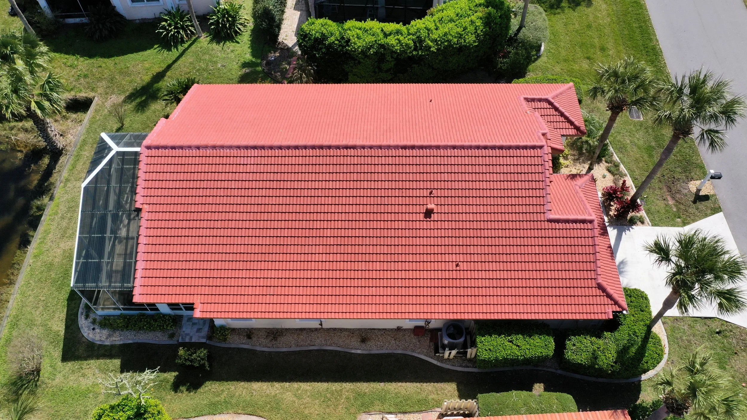 Aerial view of a house with a red tiled roof, a screened porch on the left, surrounded by green lawn and palm trees.