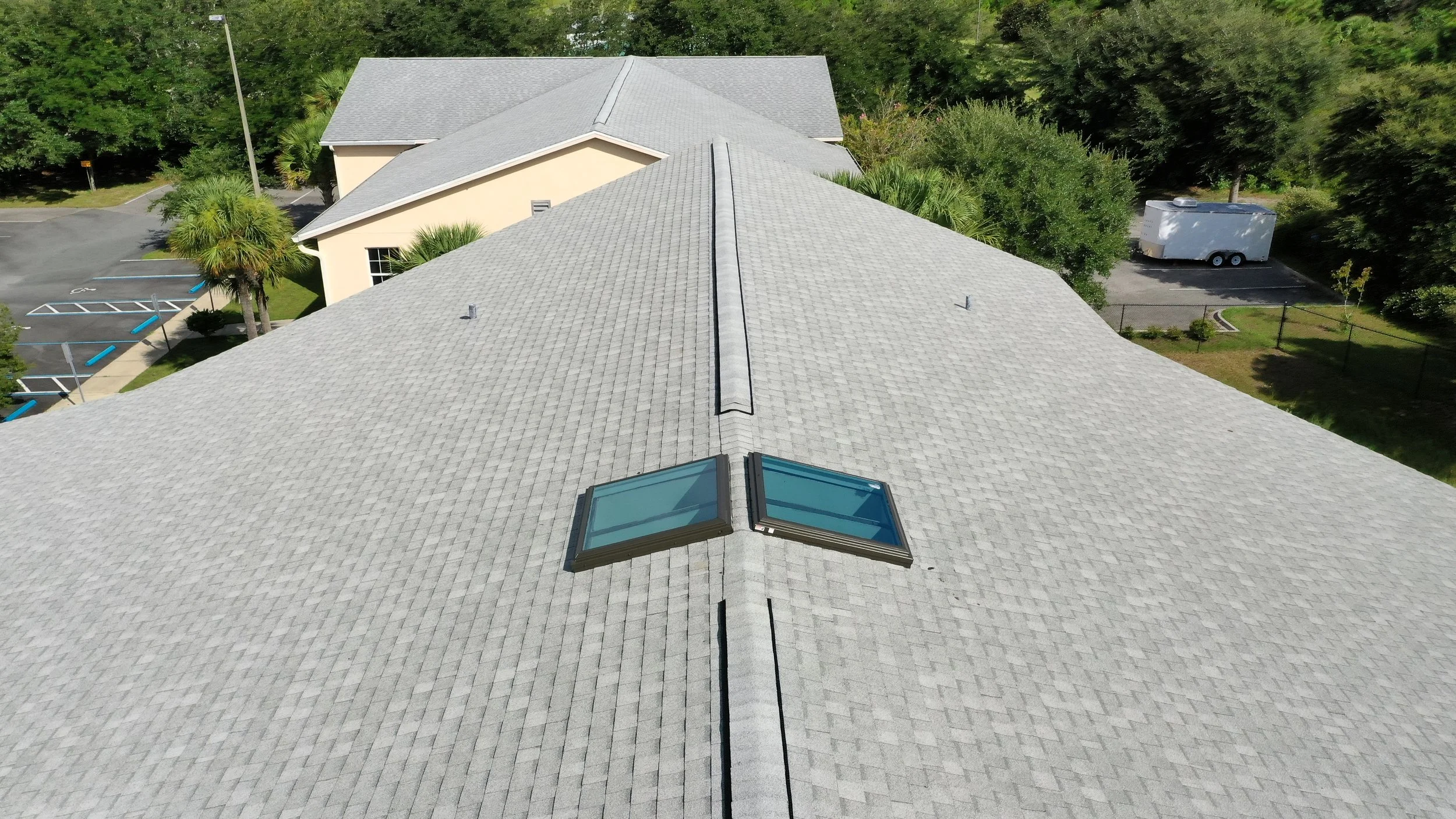 An aerial view of a light gray shingled roof with two skylights, surrounded by trees, a parking lot with a trailer, and a beige building in the background.
