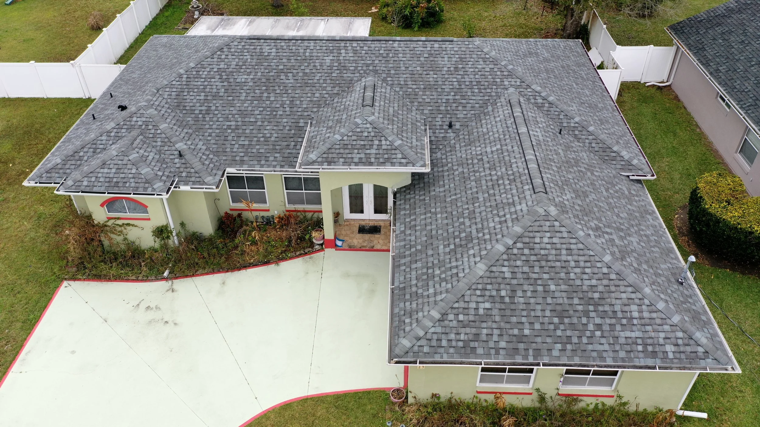 Aerial view of a house with a gray shingle roof, light green exterior walls with red trim, a small front porch, and a backyard enclosed by a white fence.