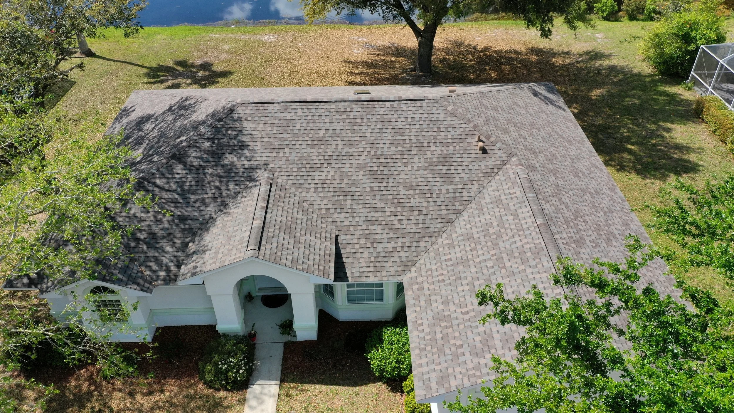 Aerial view of a house with a gray shingle roof, surrounded by green trees and backyard grass, near a lake or river.