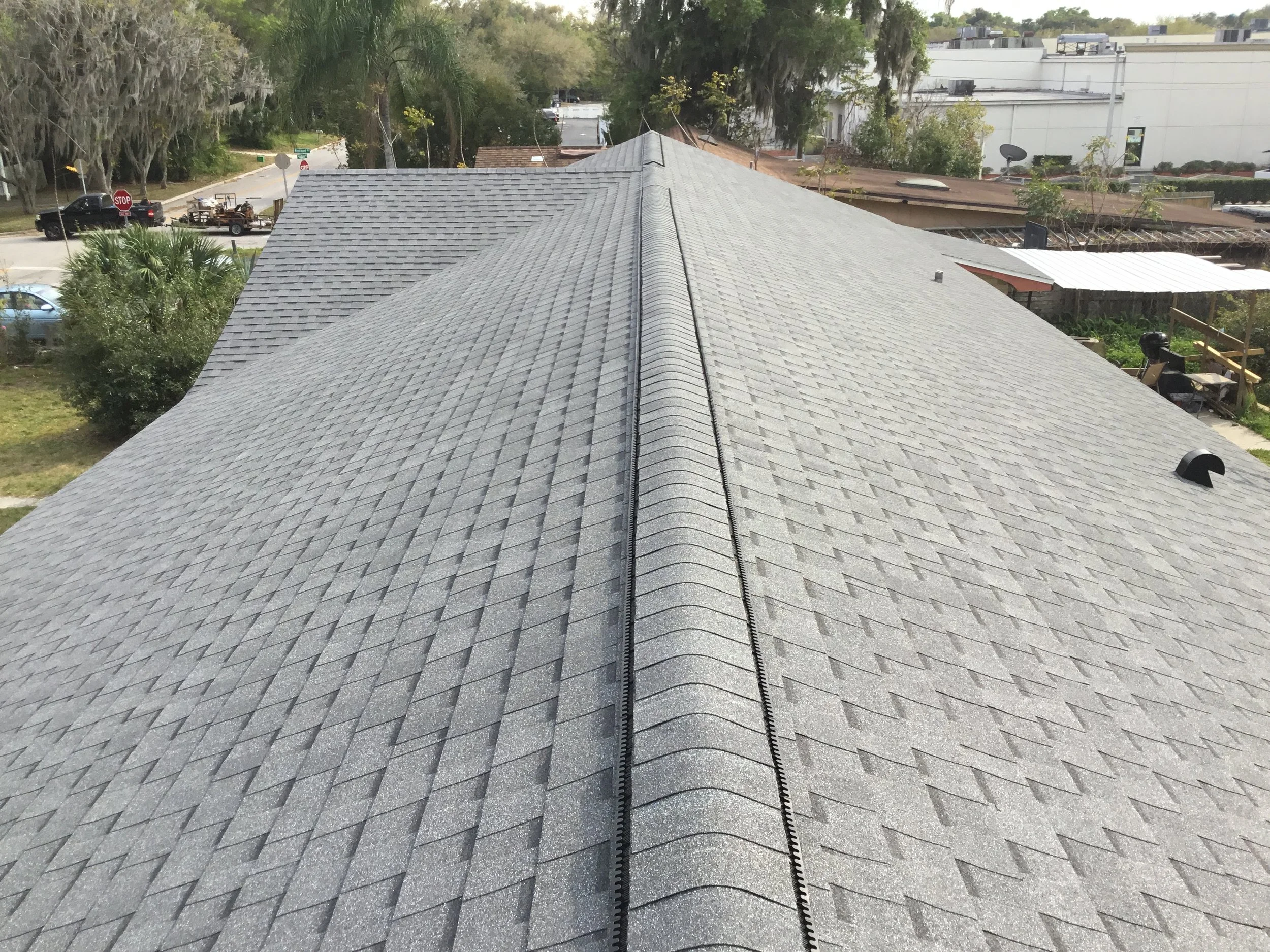 Aerial view of a house roof with gray asphalt shingles, central ridge vent, and surrounding trees and neighborhood.