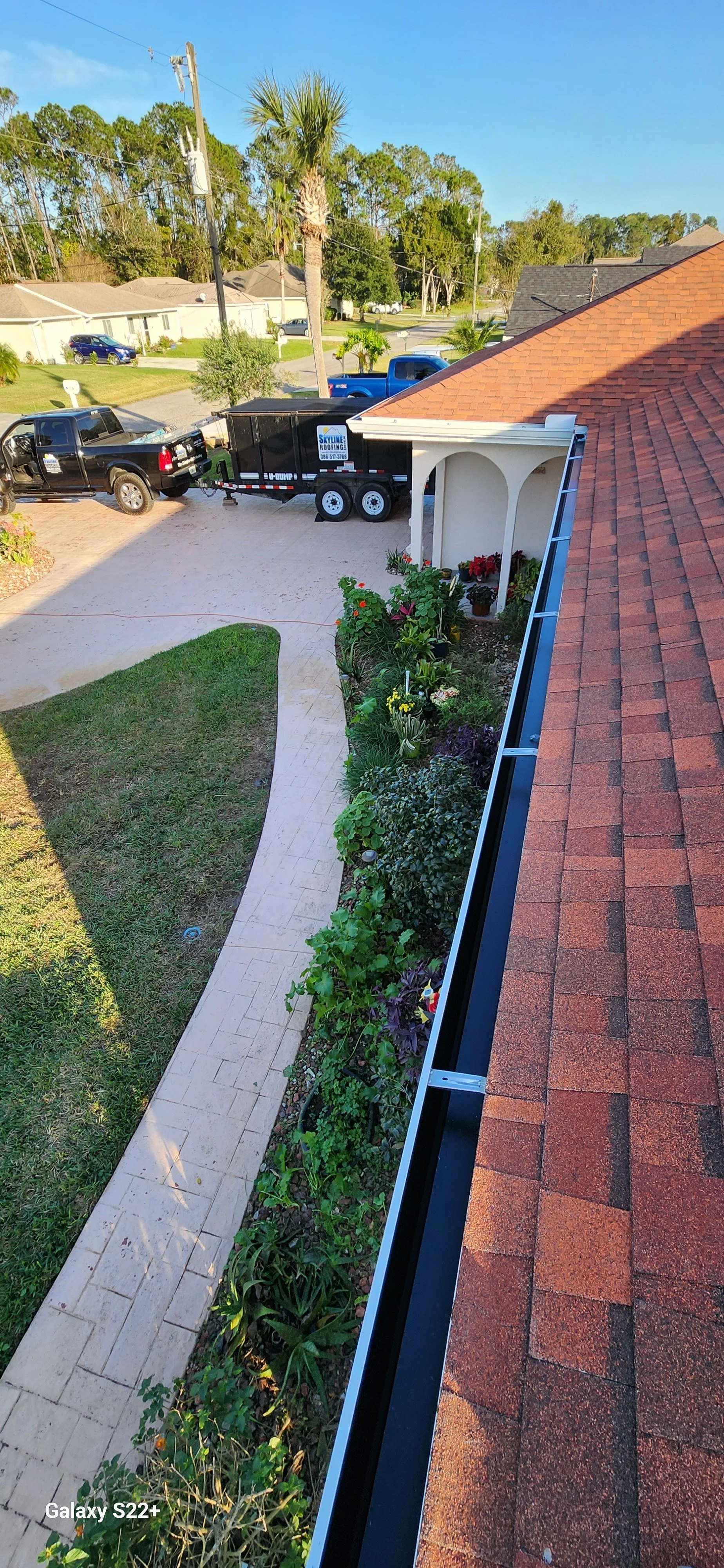 View from a house balcony showing a garden, walkway, driveway with a trailer hitched to a black pickup truck, and a neighborhood street with houses and trees in the background.