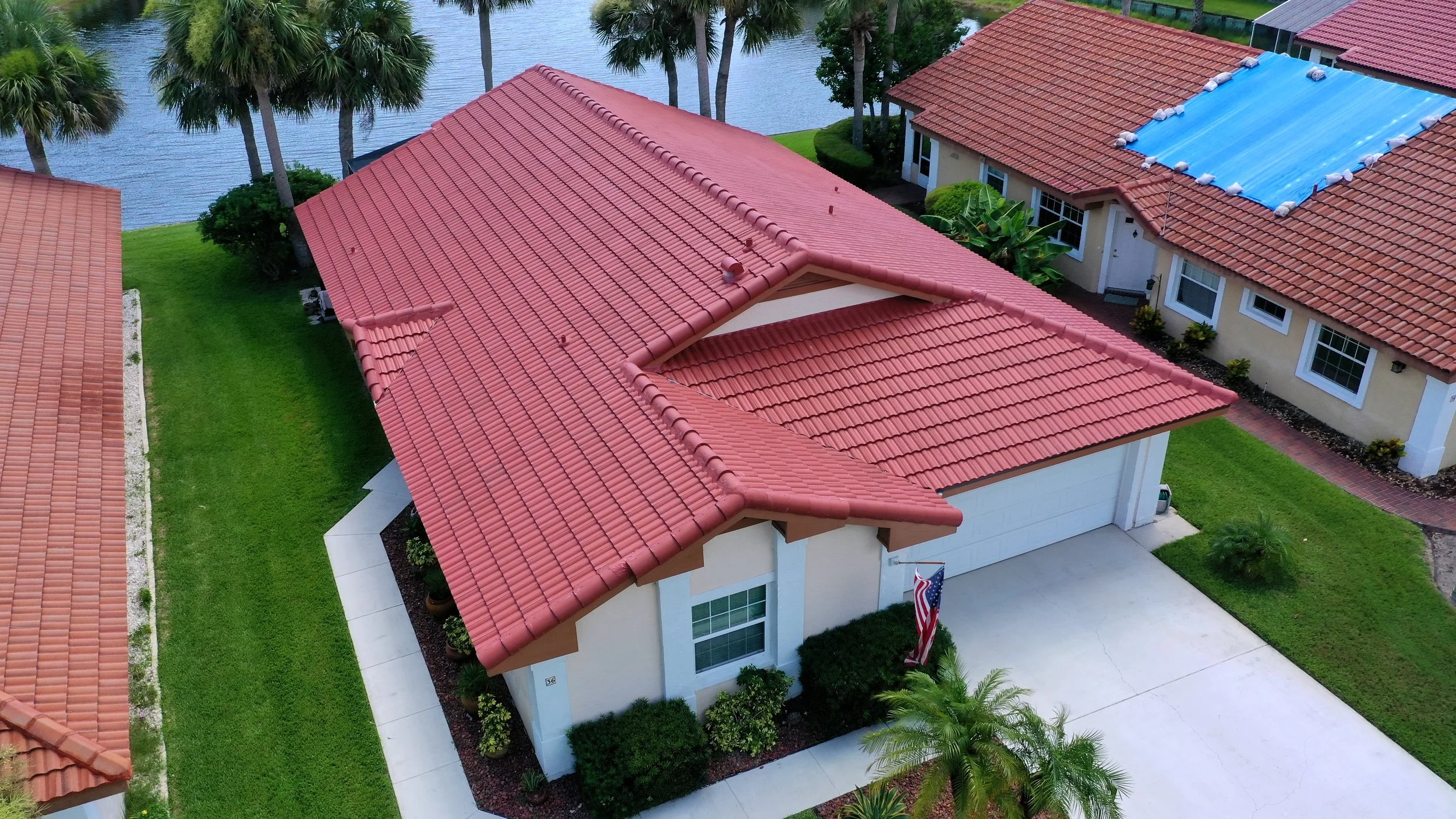 Aerial view of a house with a red tiled roof, white walls, and a attached garage. The house is surrounded by green grass and palm trees, with a body of water nearby.