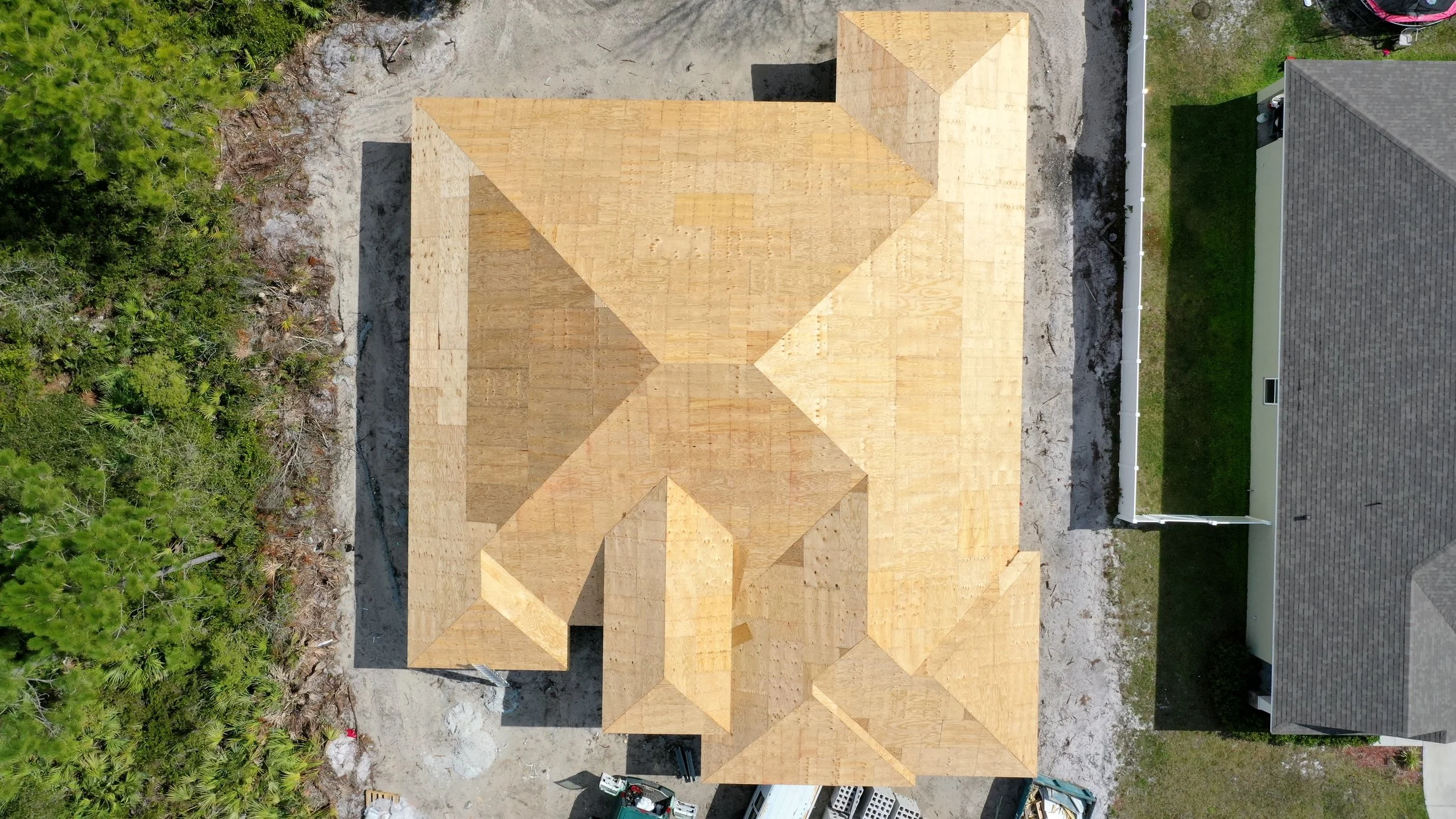 An aerial view of a house under construction with a wooden roof, surrounded by a partly developed yard and neighboring houses.
