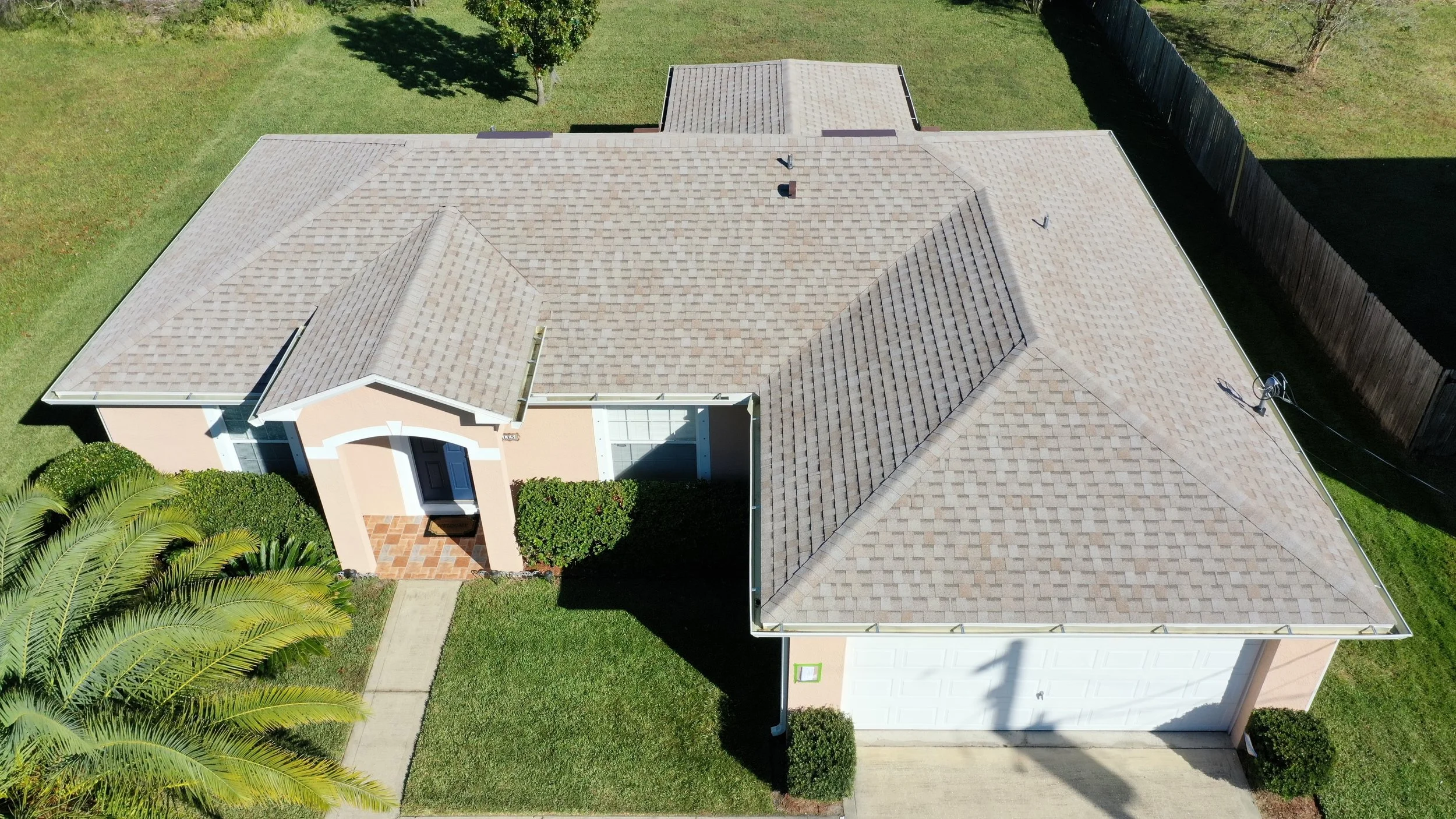 Aerial view of a suburban house with a beige roof, front entrance with a small porch, surrounded by a well-maintained lawn and trees.