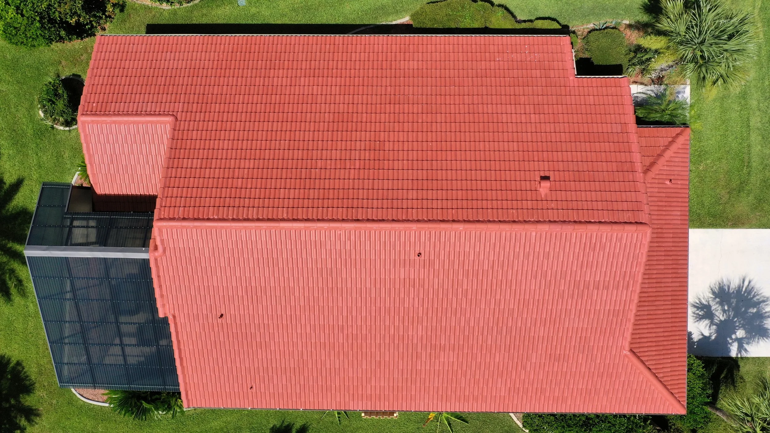 Top-down aerial view of a house with a red tiled roof and a screened-in porch extension, surrounded by green grass and tropical plants.