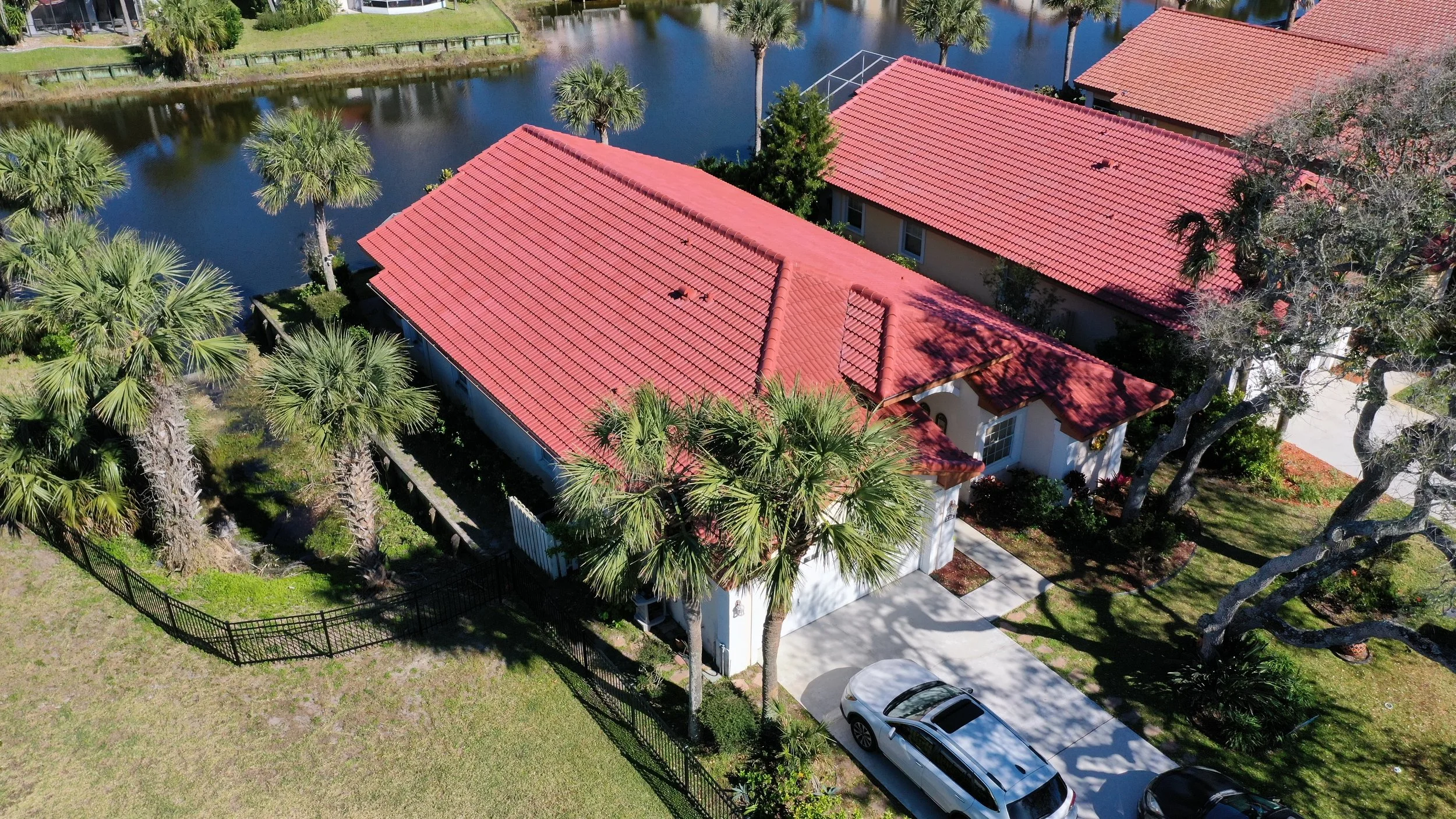 Aerial view of a house with a red tile roof, surrounded by palm trees, next to a pond with a dock, and adjacent to other similar houses.
