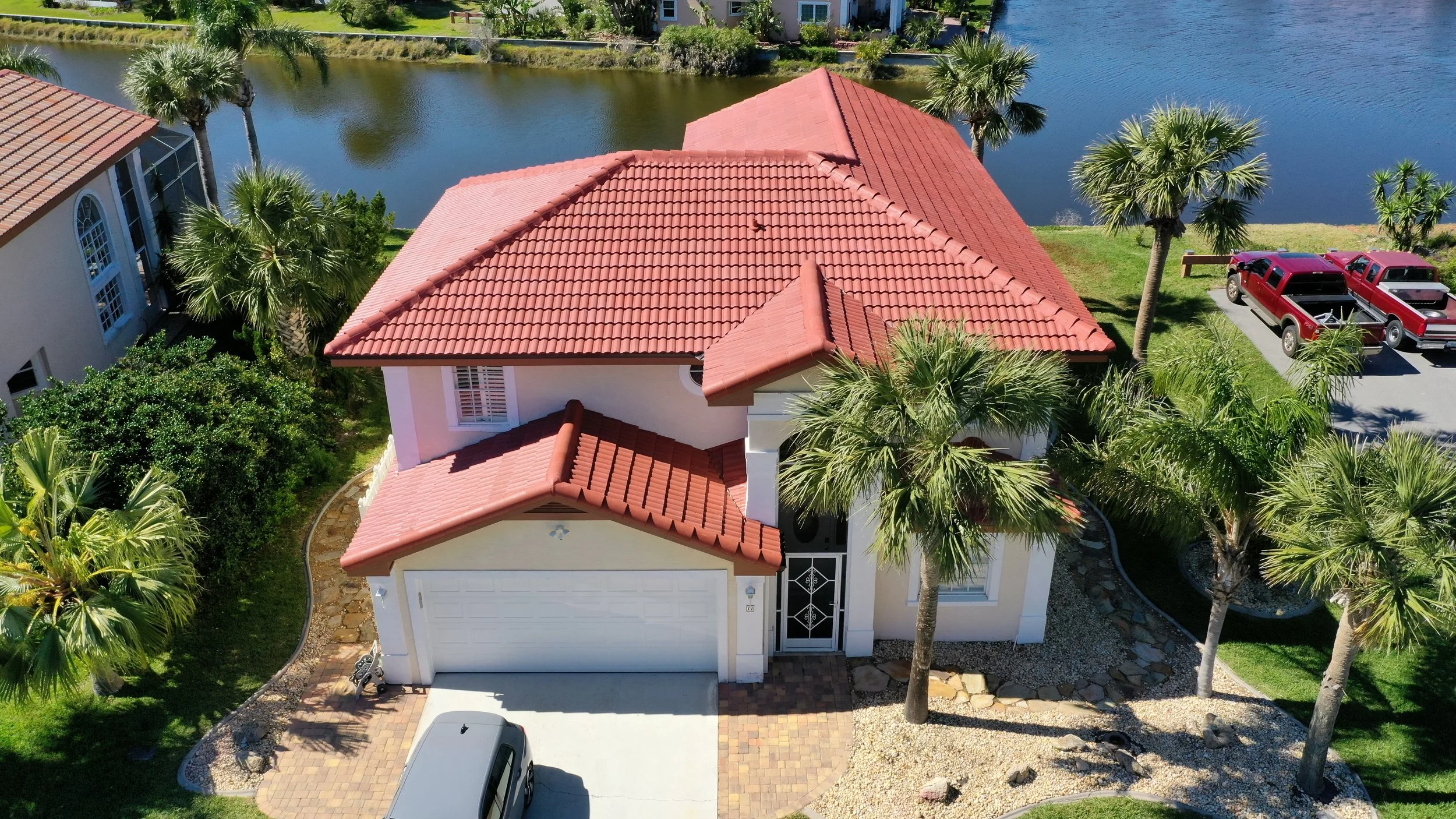 A two-story house with a red tile roof, white walls, and a garage. The house is surrounded by palm trees, bushes, and a landscaped yard. There is a paved driveway with a vehicle parked on it and a lake in the background.