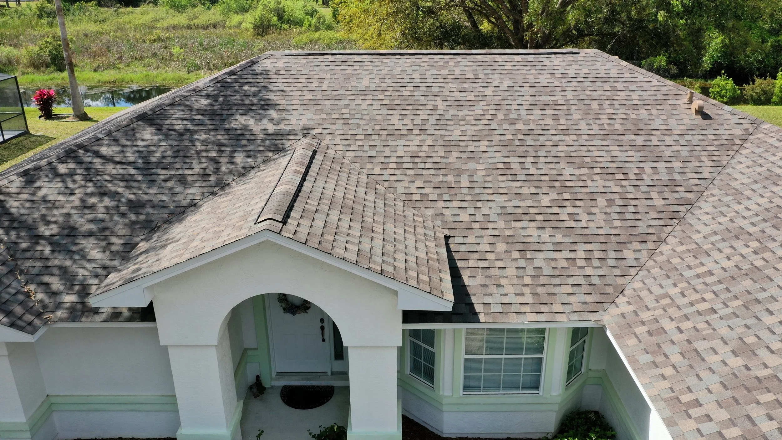 Aerial view of a house with a brown shingle roof, white exterior walls, and large windows. The house is surrounded by greenery, trees, and a small pond in the background.