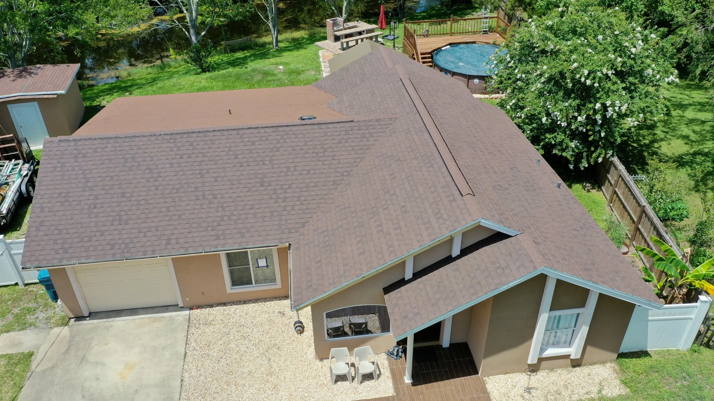 Aerial view of a residential backyard with a house, a above-ground pool, a patio area with chairs, a shed, a fenced yard with trees, and a gravel driveway.