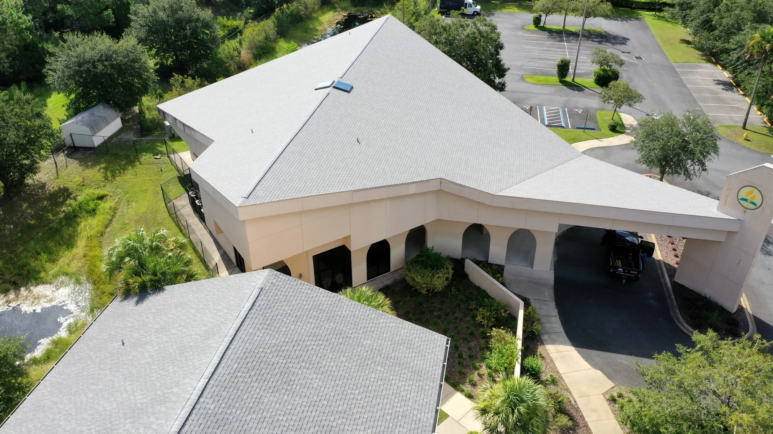 Aerial view of a building with a gray roof, a driveway with a black truck, surrounded by trees, a parking lot, and a small pond.