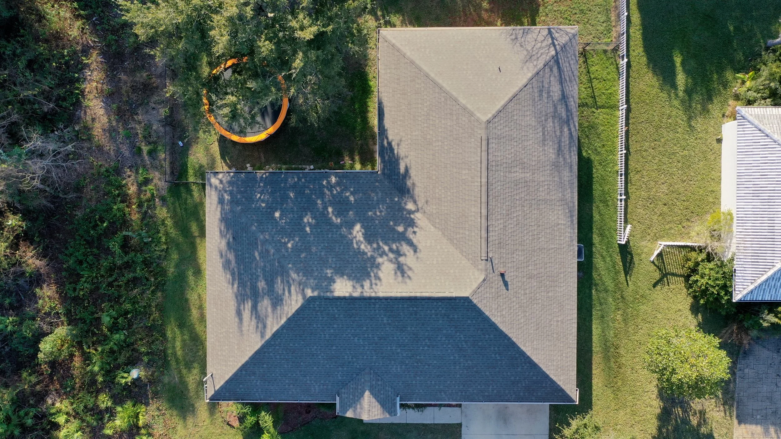 Aerial view of a house with a grey shingled roof, surrounded by green lawn, trees, and neighboring houses. A trampoline is visible in the backyard. Shadows of trees are cast on the roof.